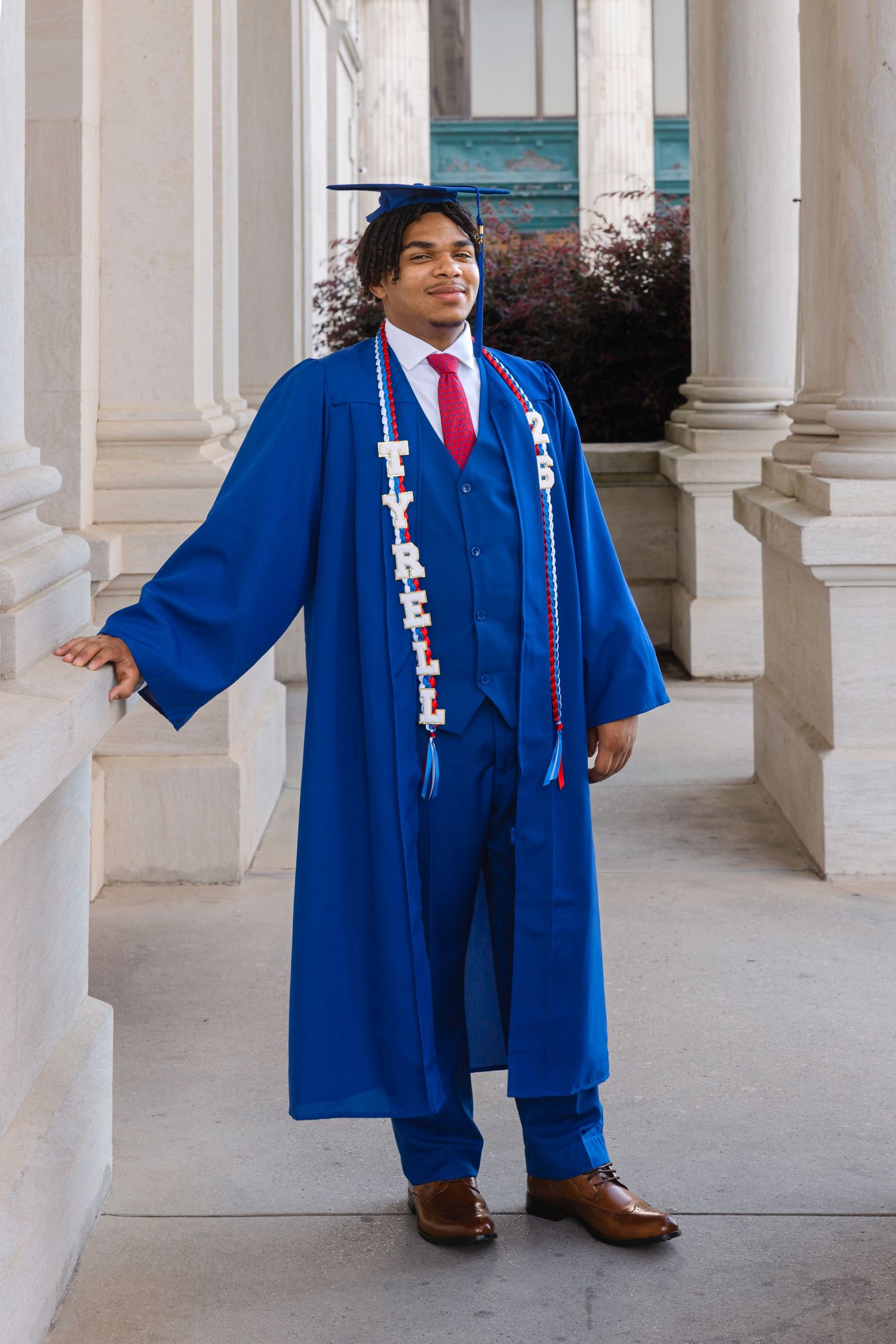 Young man in blue graduation cap and gown with name sash poses between white columns at commencement.