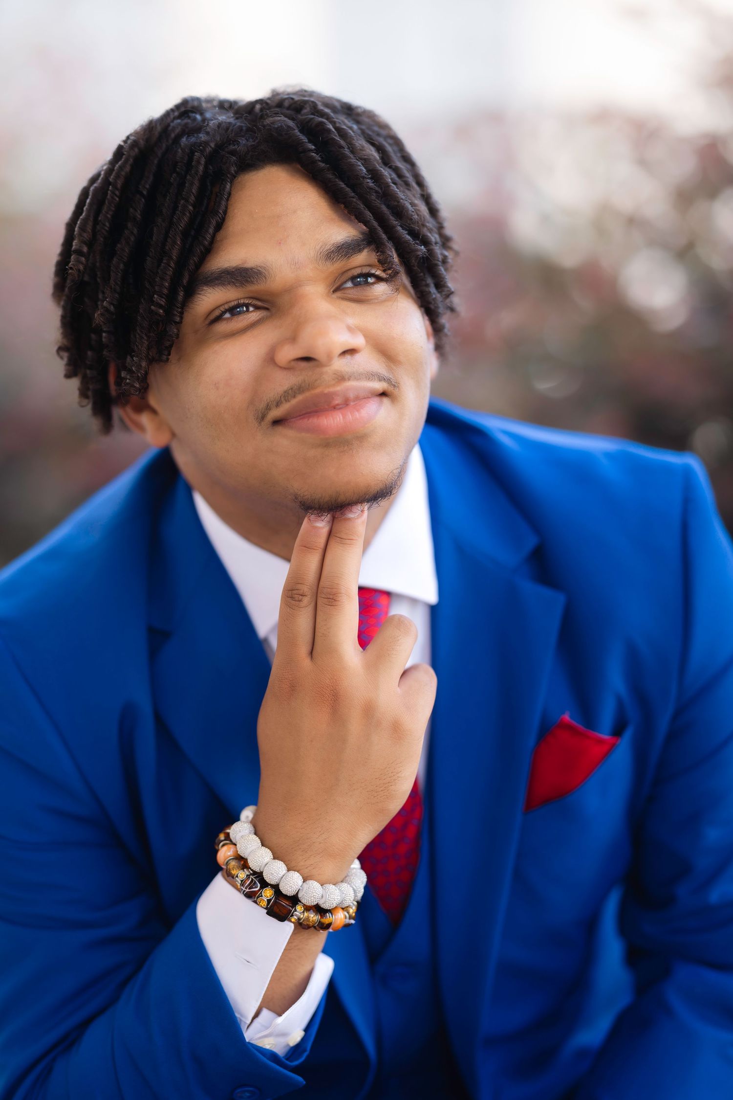 Young man in blue suit with red tie and pocket square, wearing beaded bracelets, posing thoughtfully outdoors.