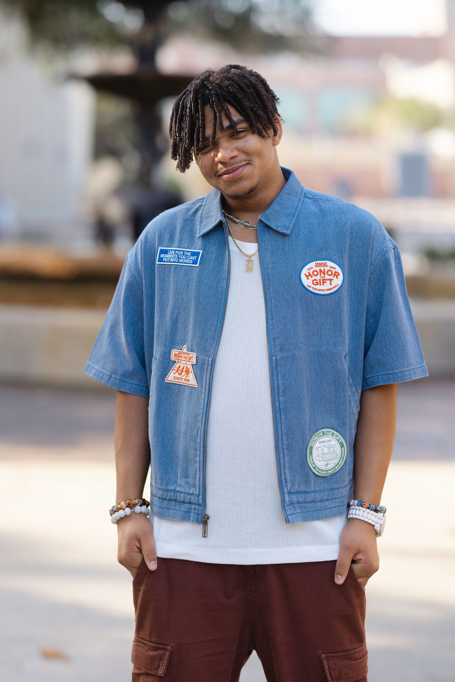 Young man in denim patch shirt and cargo pants smiling outdoors near a fountain.