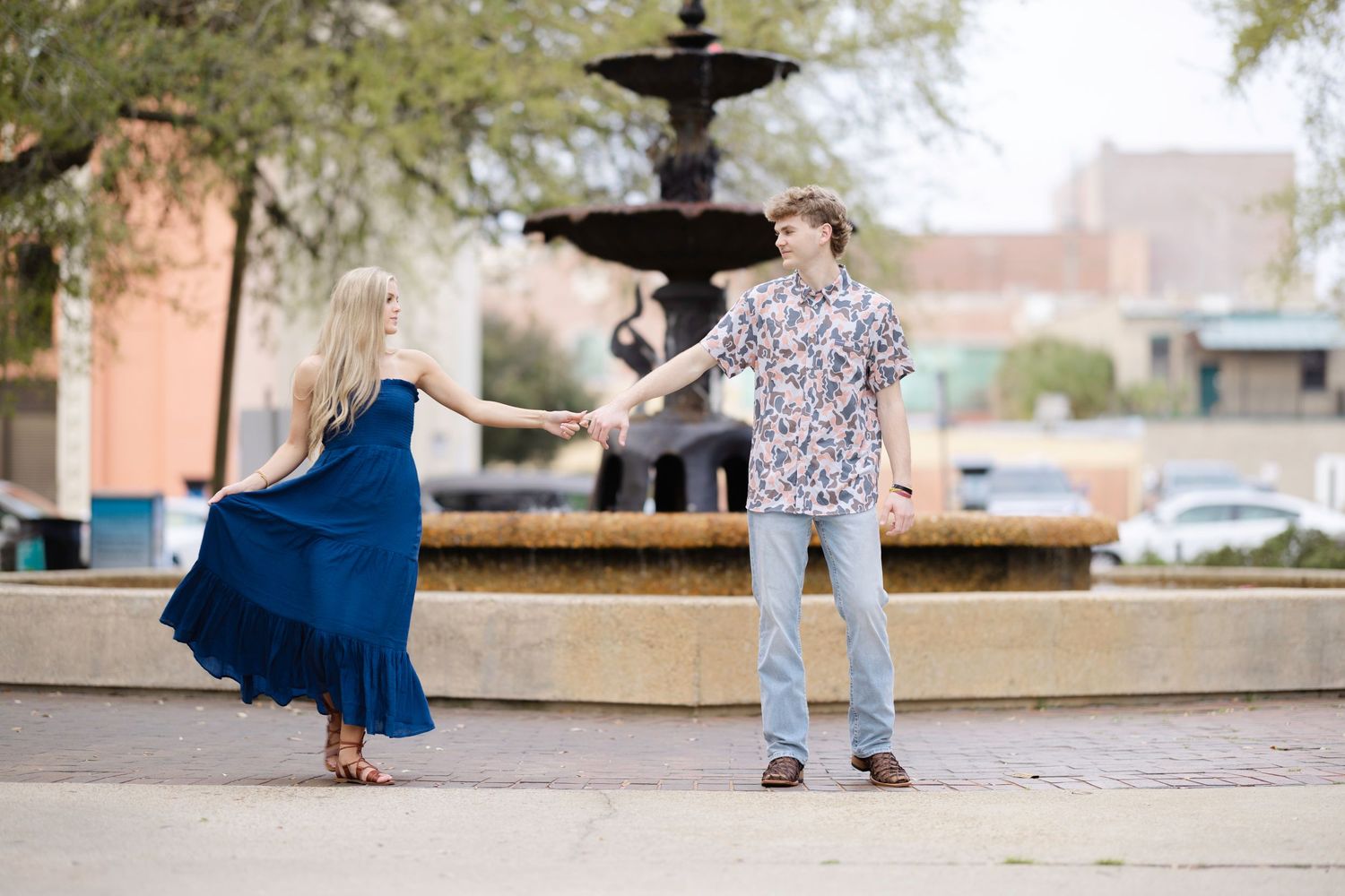 Two people dance together in front of a fountain in an outdoor plaza, with one wearing a blue dress and one in casual attire.