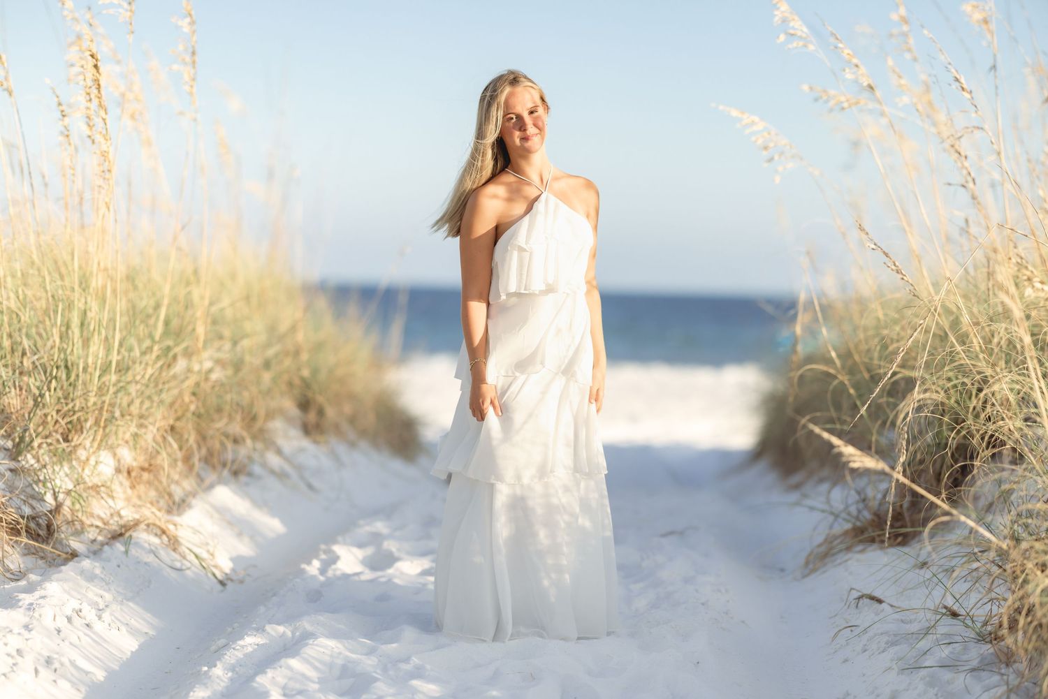 Blonde woman in white maxi dress standing on sandy beach path with sea oats and ocean in background.