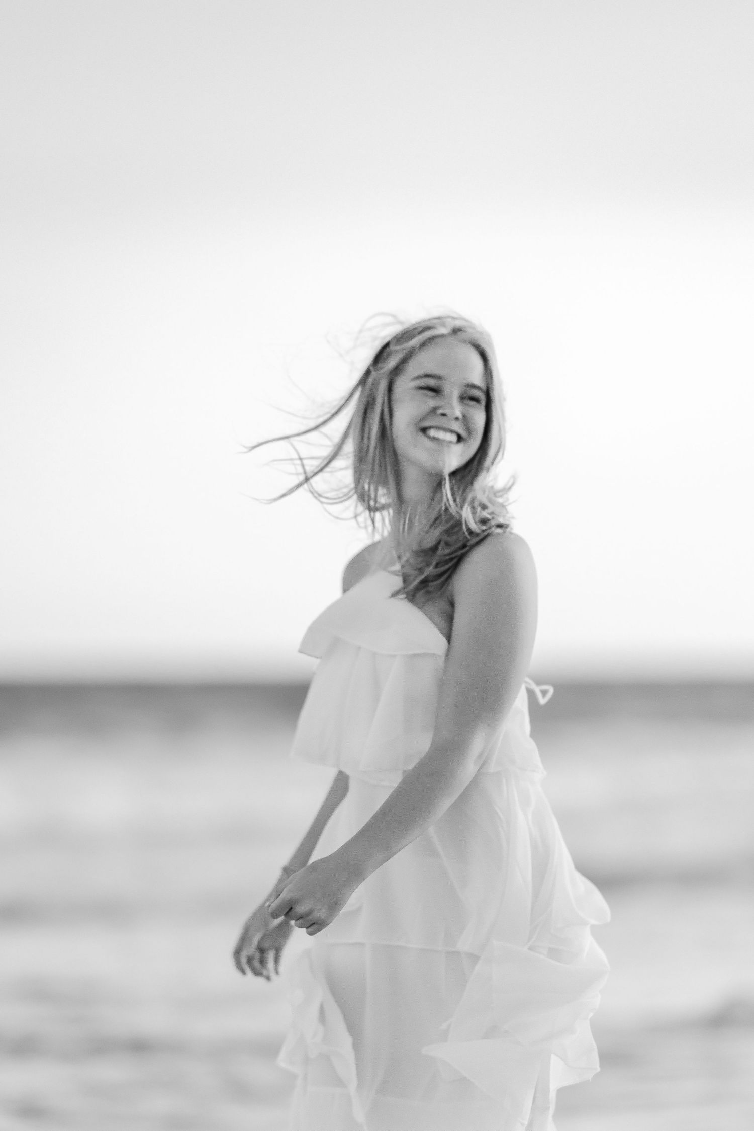 Black and white photo of a smiling young woman in a white dress on a beach with wind blowing her hair.