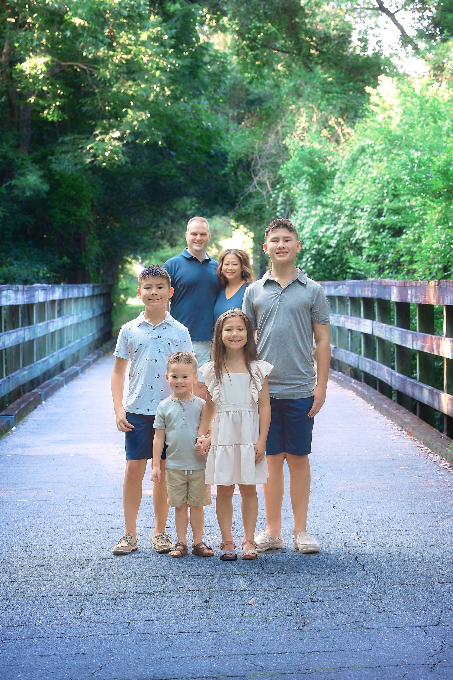 Family of six posing on a wooden bridge surrounded by lush green trees during an outdoor portrait session.