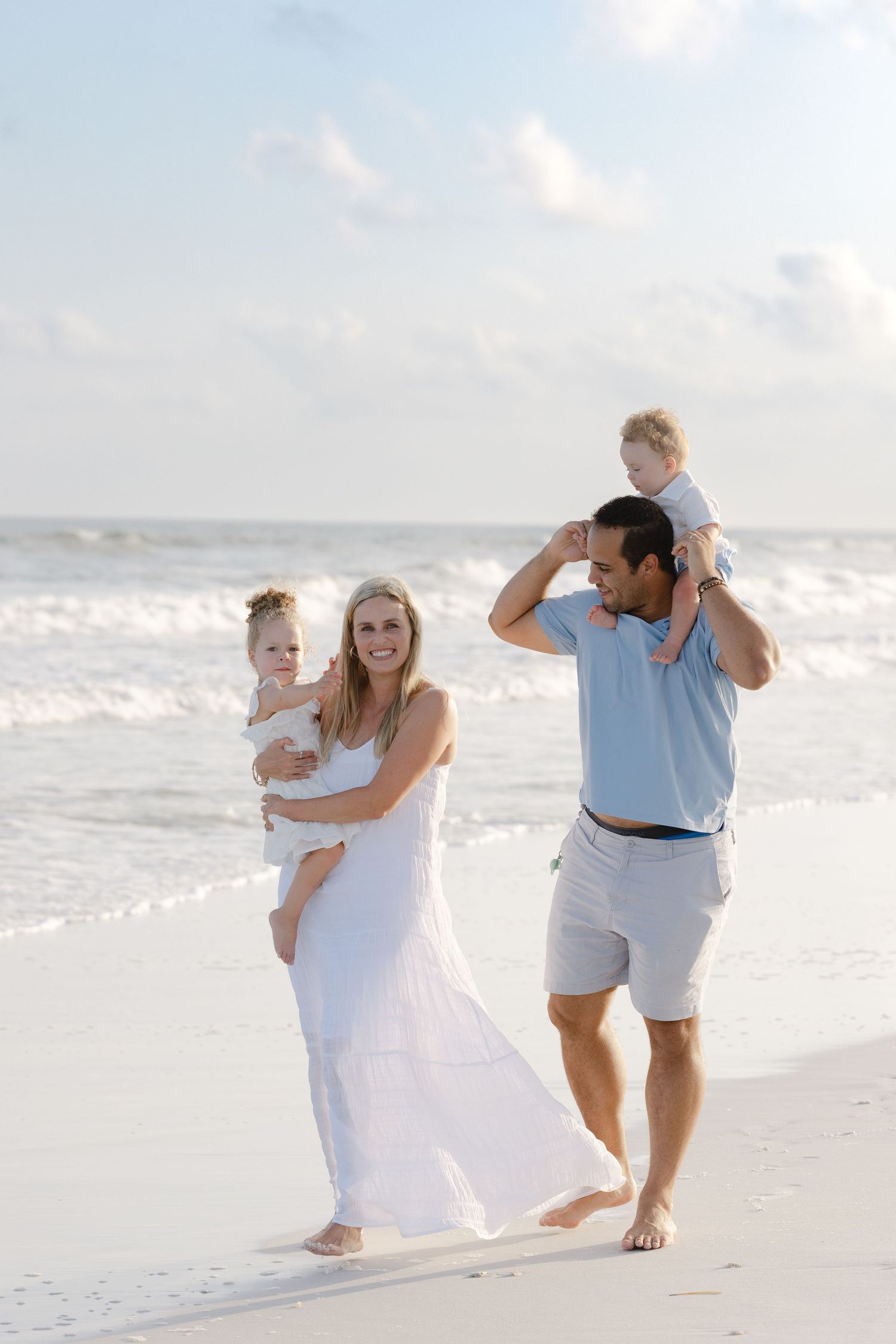 Happy family with two young children enjoying a sunny day on the beach, mother in white dress holding baby.