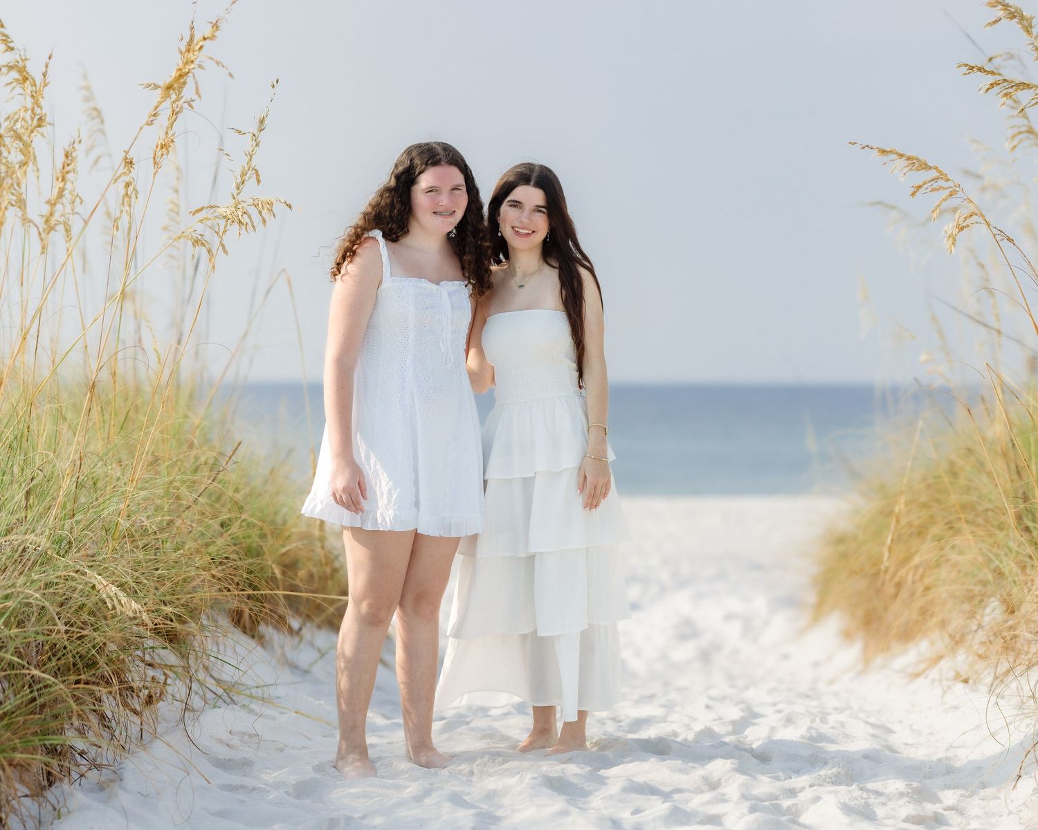 Two young women in white outfits smiling together on a sandy beach with sea oats.