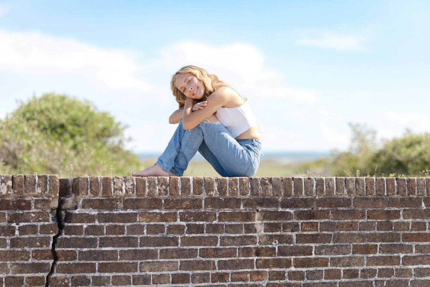 A figure seated on a brick wall wearing jeans gazes into the distance as sunlight illuminates the scene.