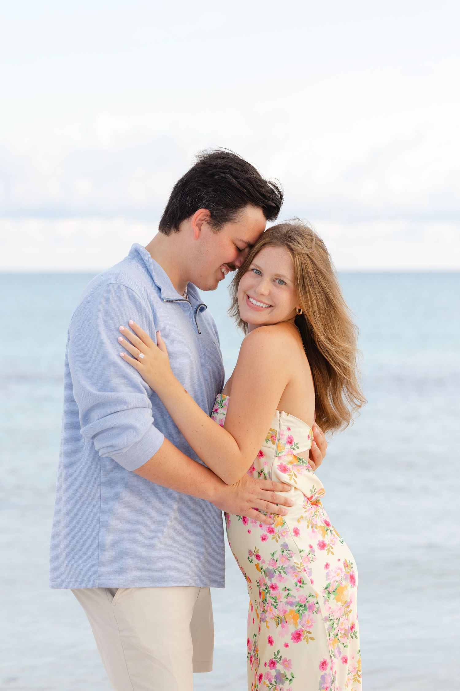 Romantic couple embracing on beach with ocean view, wearing floral dress and blue shirt.