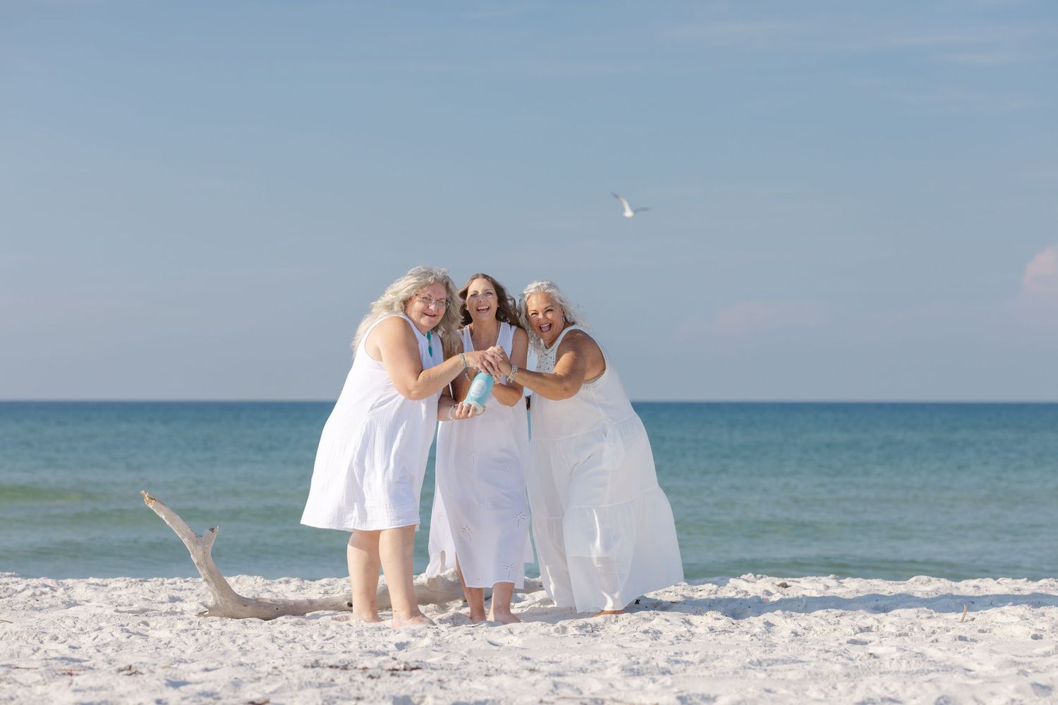Three people in white dresses embrace while standing on a sunny beach with ocean waves in the background.