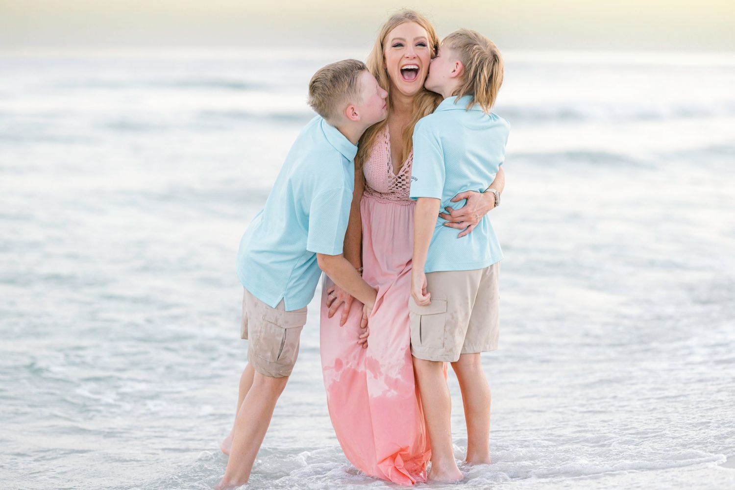 Two people in pink and blue outfits share a romantic moment on the beach at sunset near the ocean waves.