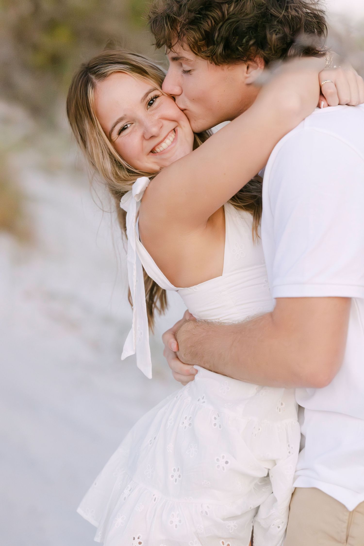 A couple in white outfits share an intimate embrace and kiss outdoors in soft lighting.