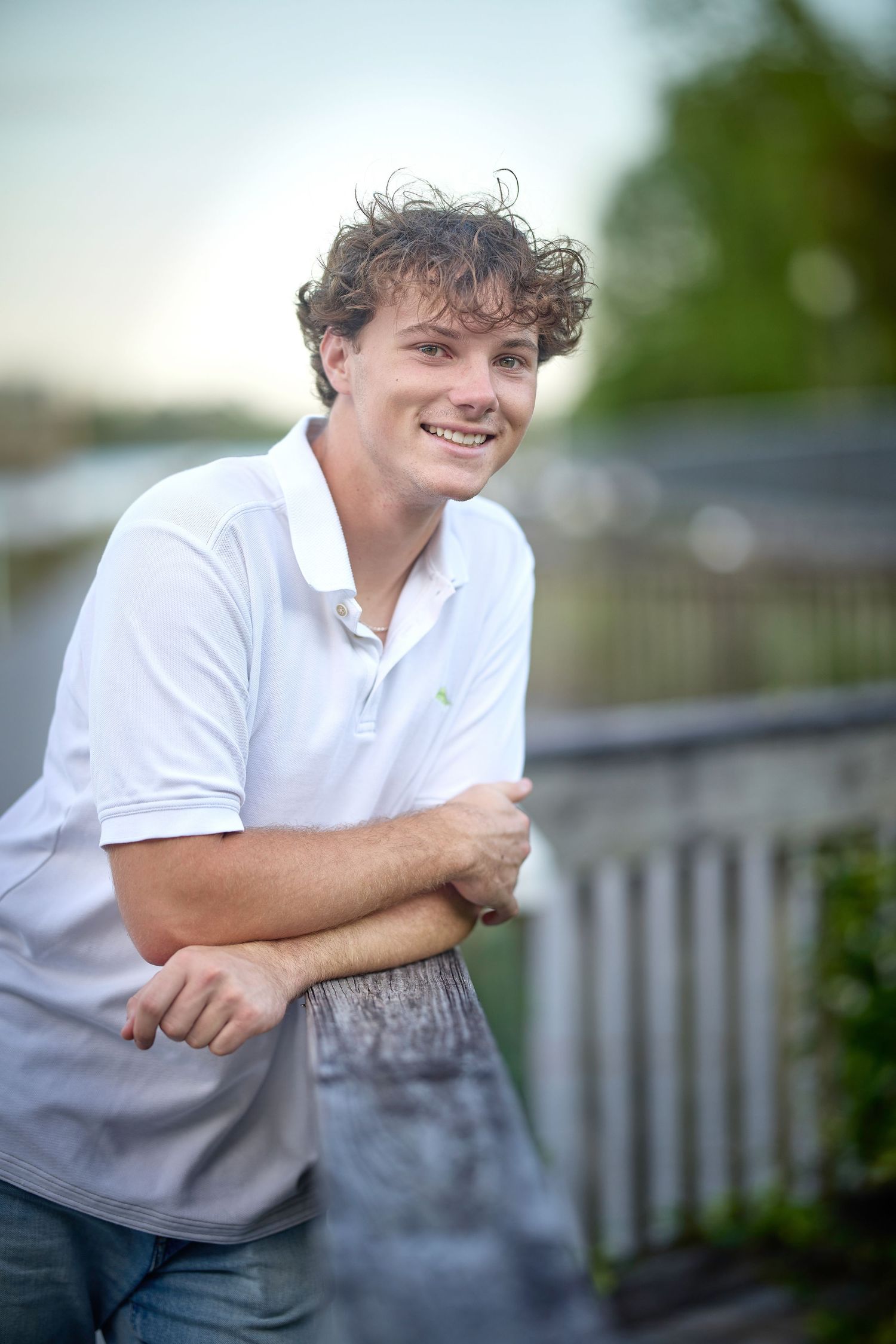 A person in a white polo shirt leans against a railing outdoors, with a soft blurred background.
