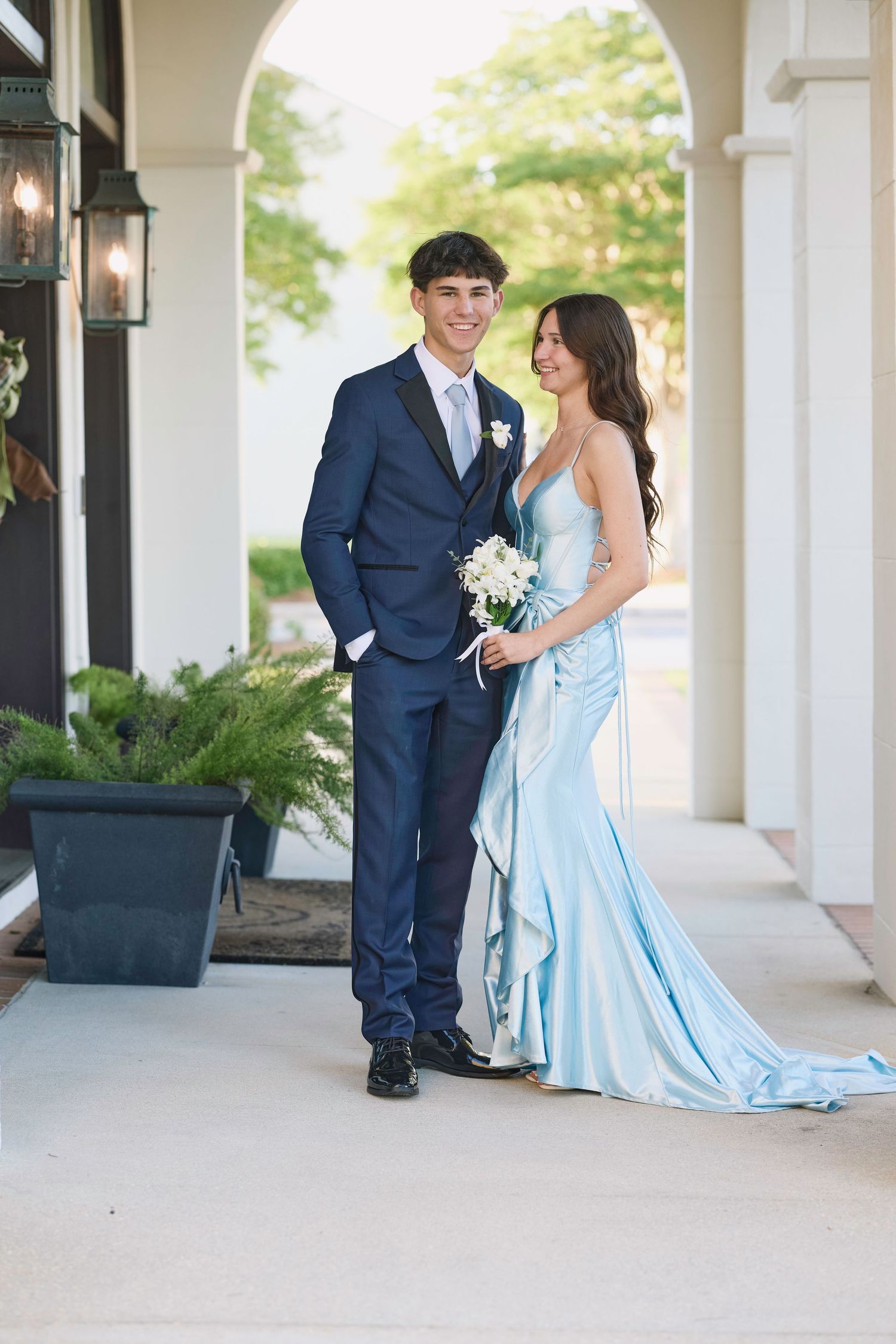 A couple poses under an arched entryway, with the woman wearing a flowing light blue gown and holding a white bouquet.