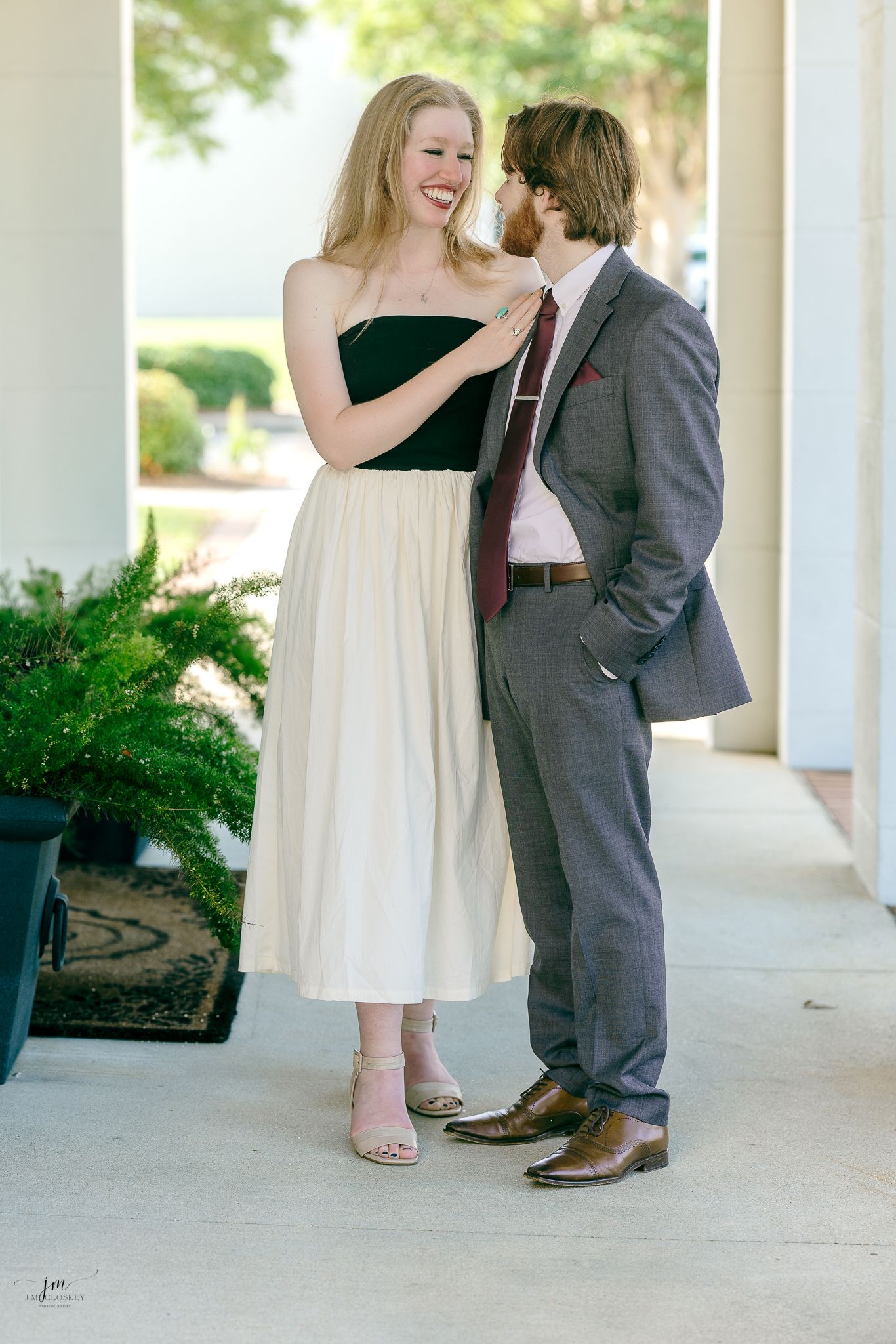 A couple shares a joyful moment together on a covered porch, with her in a cream dress and him in a grey suit.