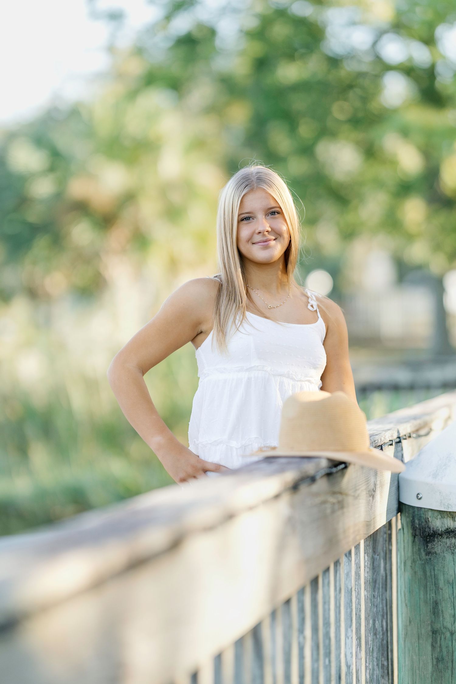 Person in a white sleeveless top poses near a wooden railing with a straw hat on a sunny summer day.