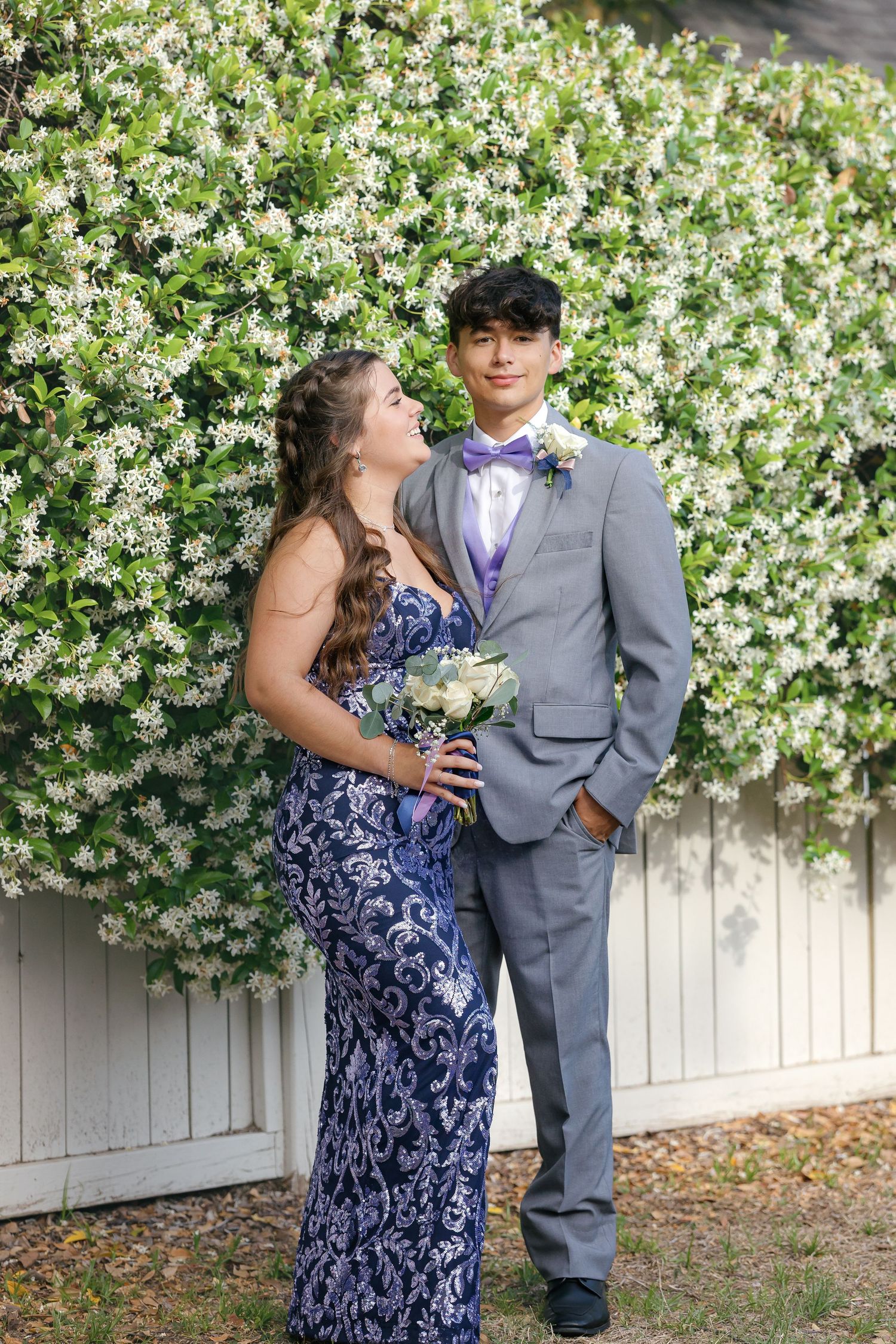 Young couple in formal attire pose for prom photo against white flowering wall in garden setting.