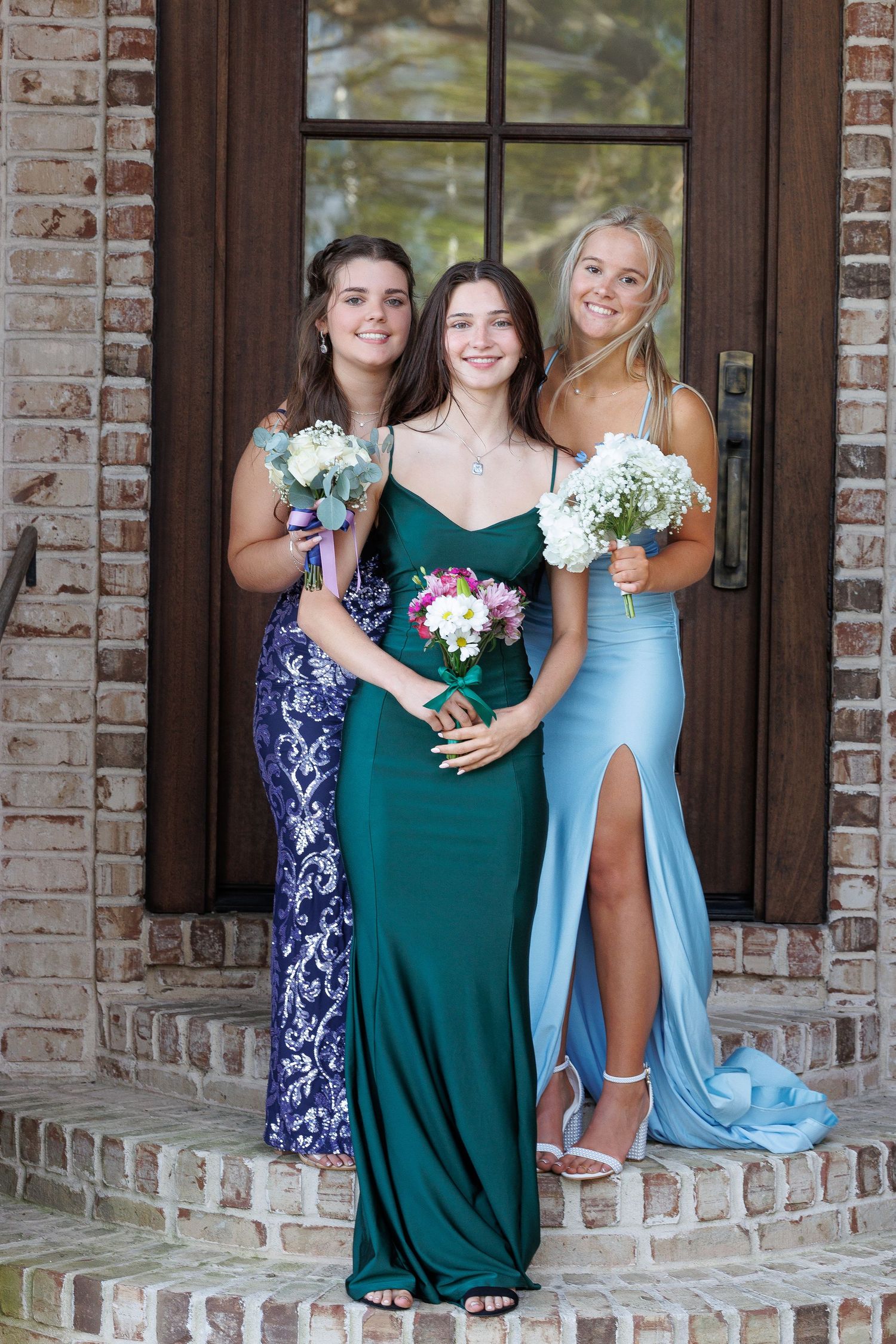 Three women in formal dresses holding floral bouquets pose on brick steps in front of a dark wooden door.