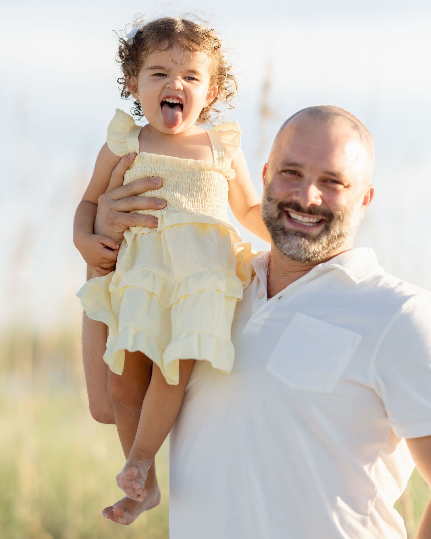 A smiling bald man with a beard holds a laughing toddler girl in a yellow ruffled dress outdoors.