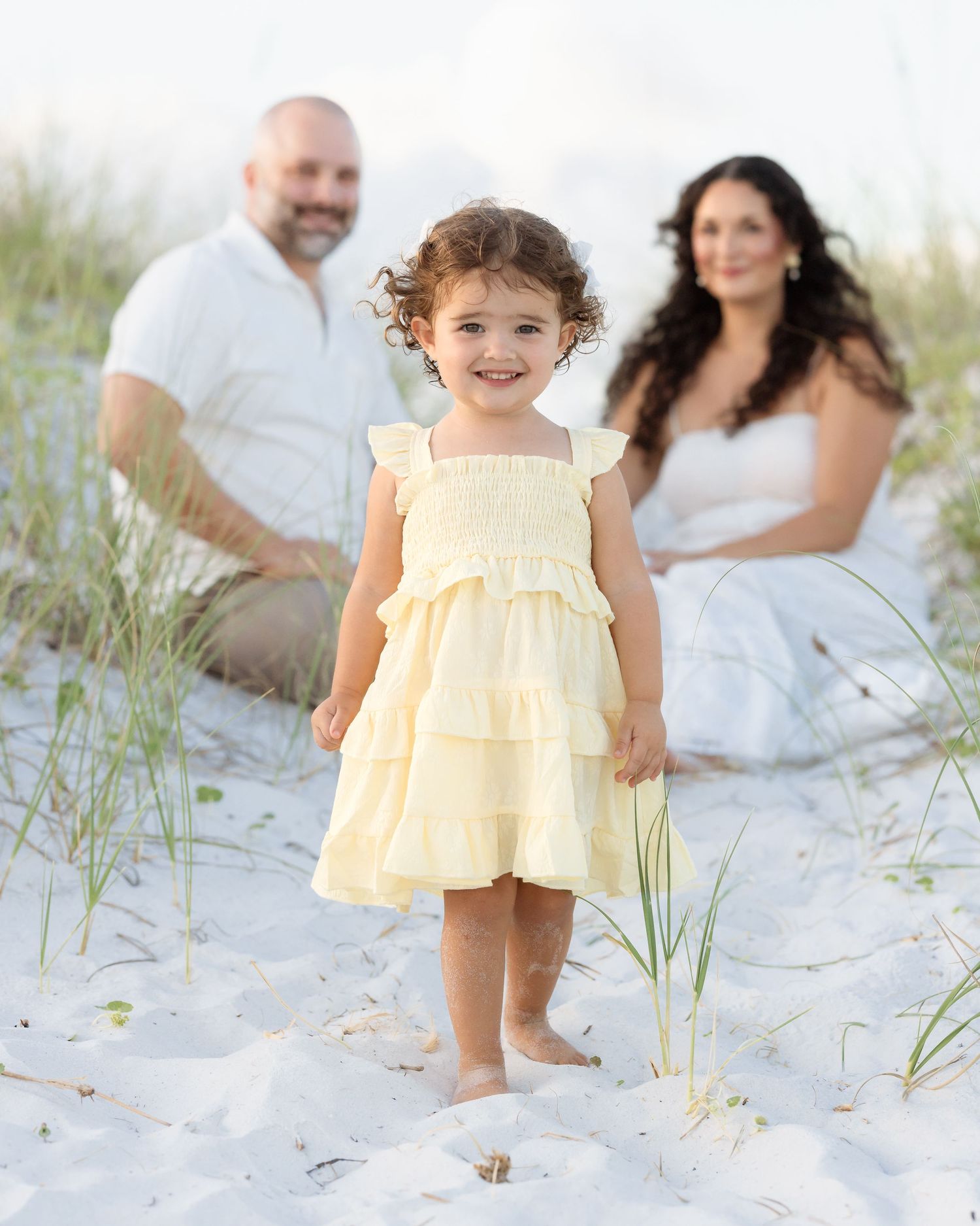 Young family with toddler girl in yellow dress posing in white sand beach dunes.