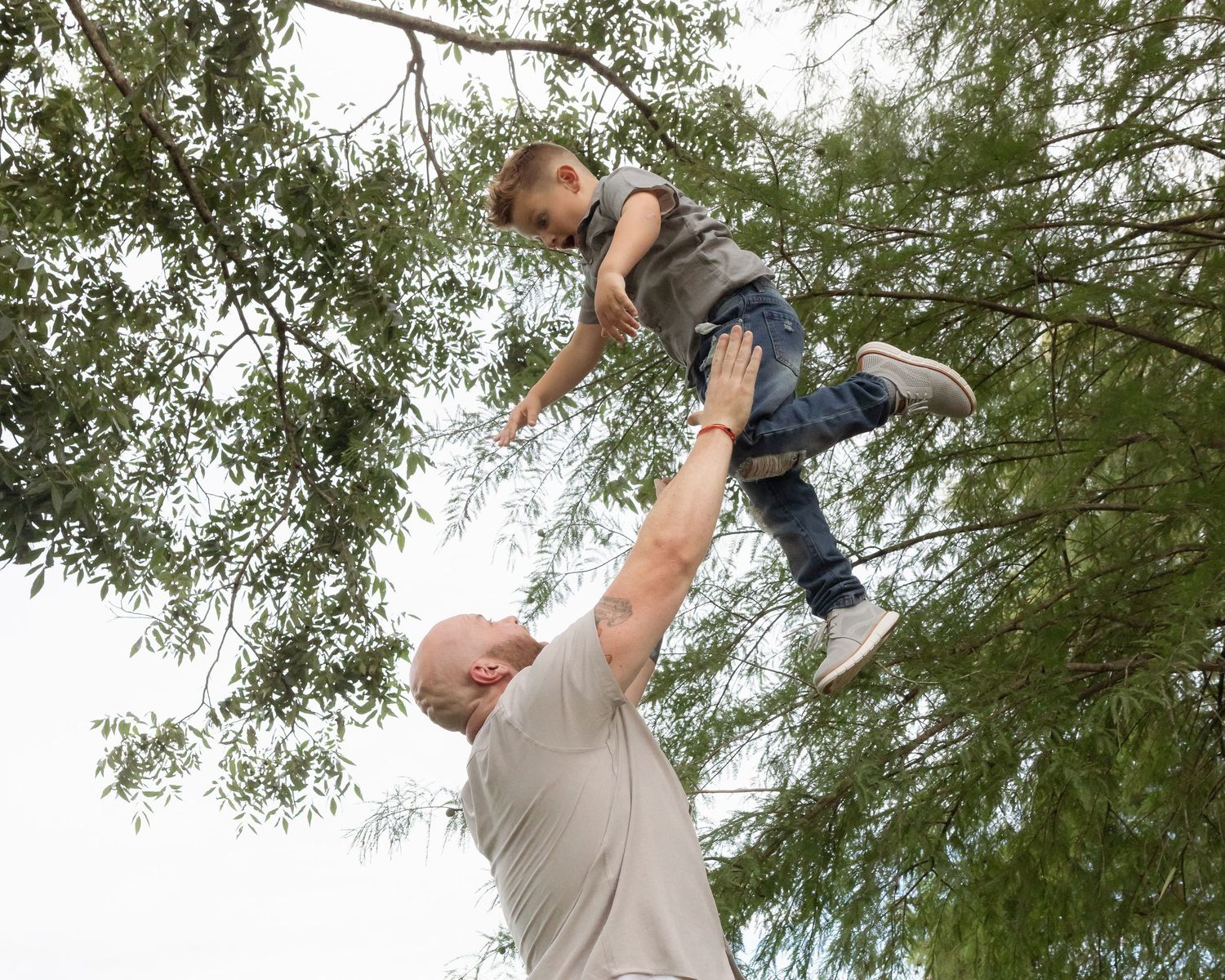 Father lifting young child high into the air outdoors beneath green tree branches.