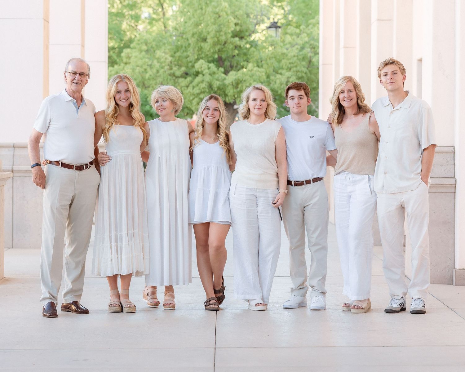 A family of eight poses together in coordinating white and tan outfits outdoors near elegant columns.