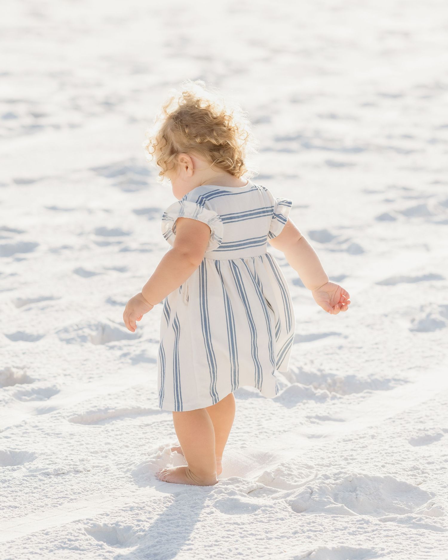A toddler girl in a white and blue striped dress walks barefoot on white sand beach.
