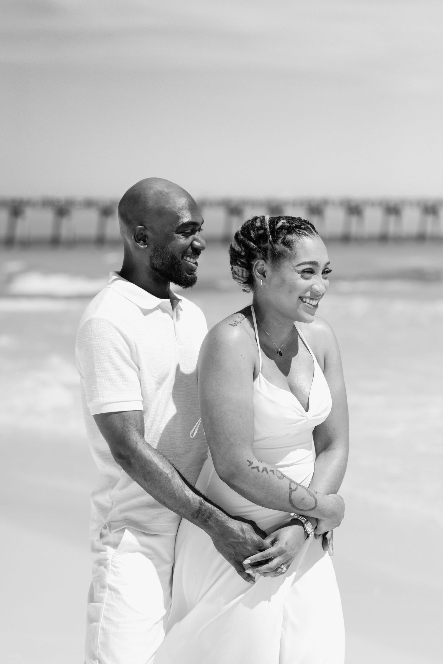 Black and white photo of a smiling couple in white attire embracing on a sunny beach with a pier in the background.