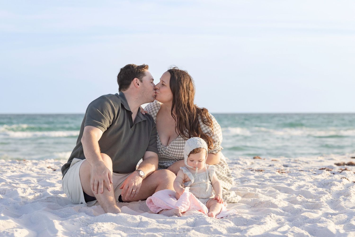 A couple kissing on a white sand beach while their baby sits beside them with ocean waves in the background.
