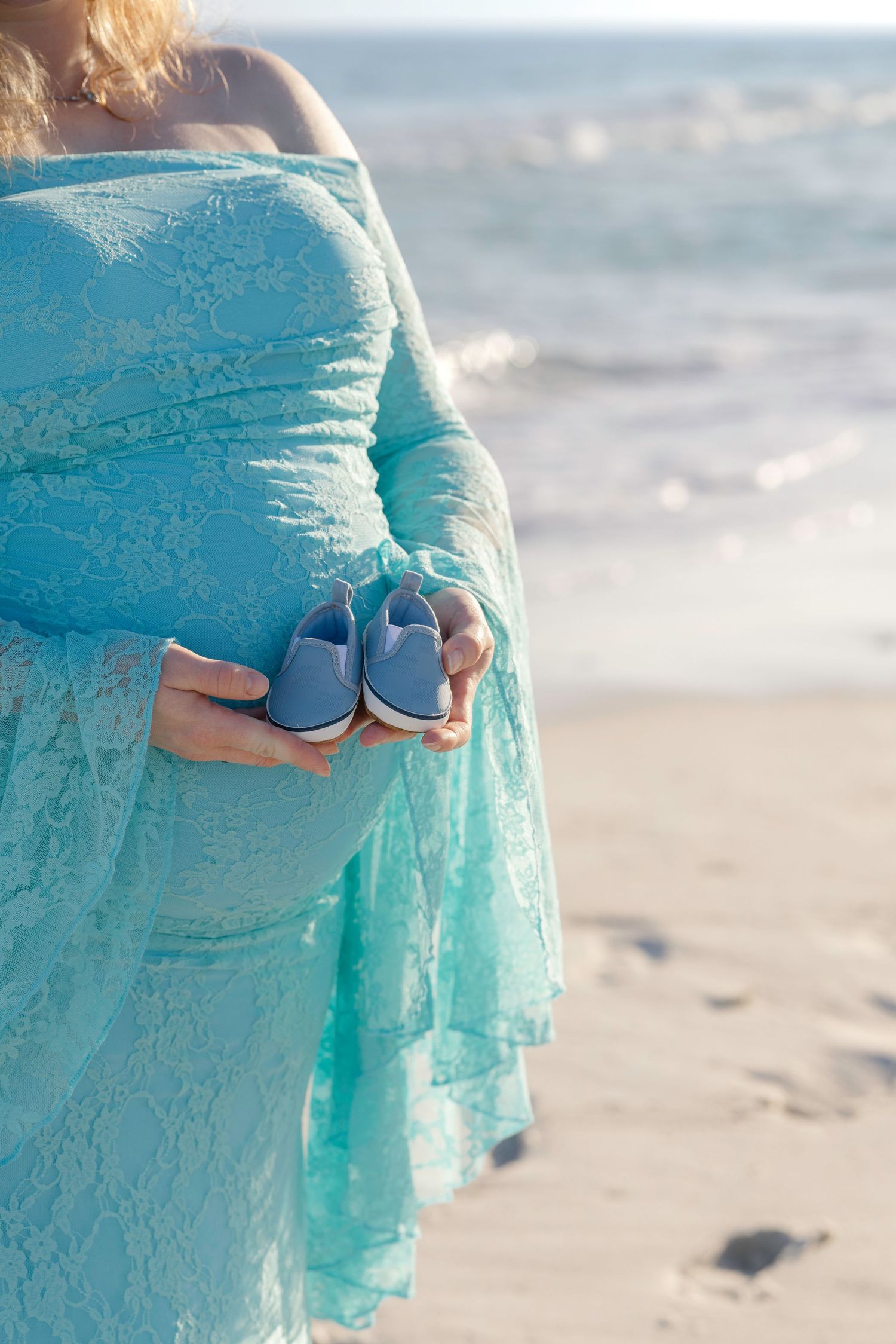 Pregnant woman in teal lace dress holding tiny blue baby shoes on a sunny beach.
