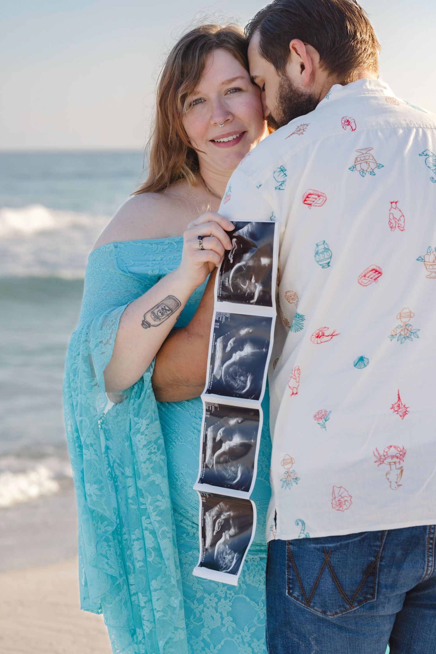 Pregnant couple on beach holding ultrasound photos, woman in teal lace dress smiling, man kissing her cheek at sunset.