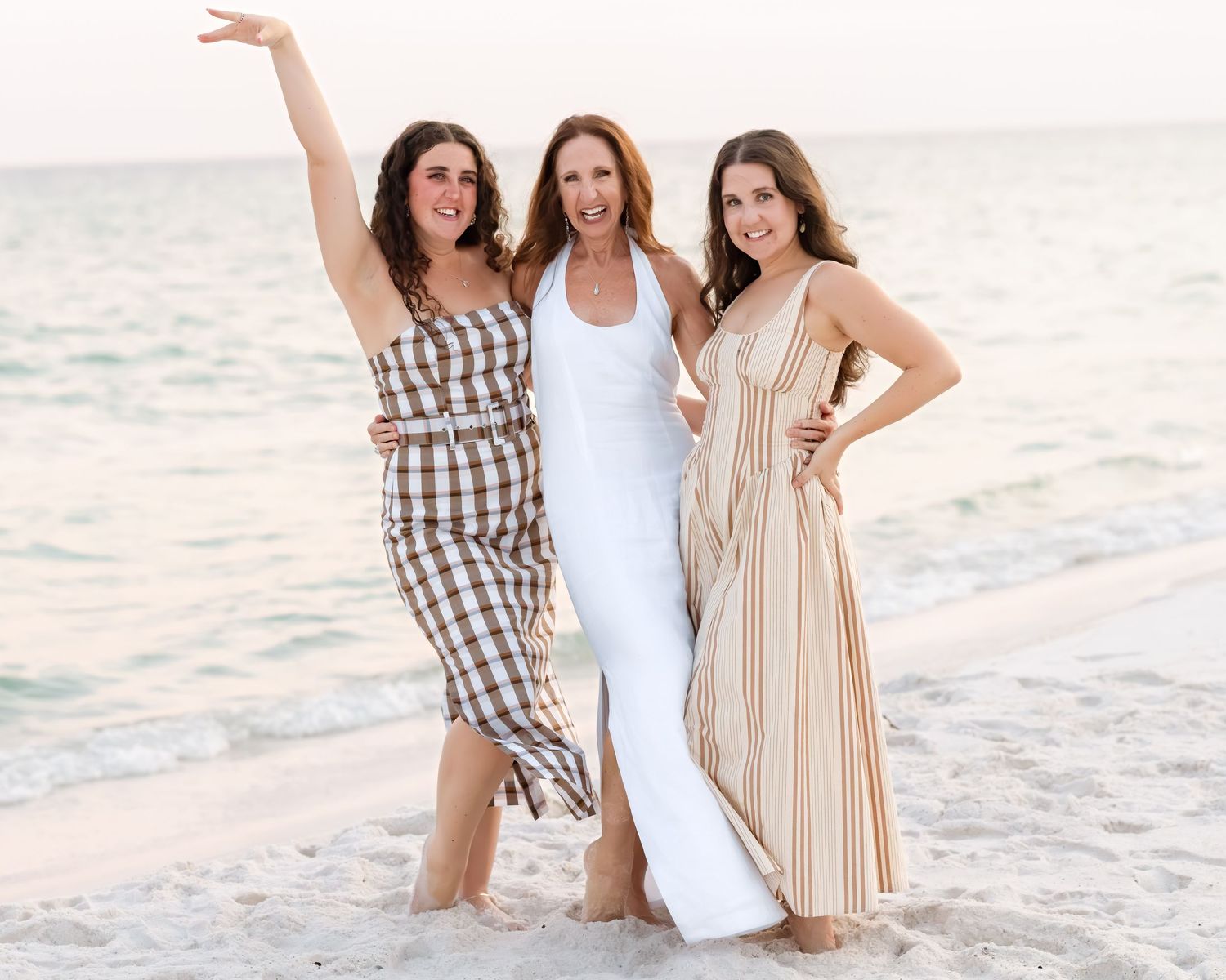 Three women smiling and posing together on a sandy beach, wearing summer dresses in white, plaid, and striped patterns.