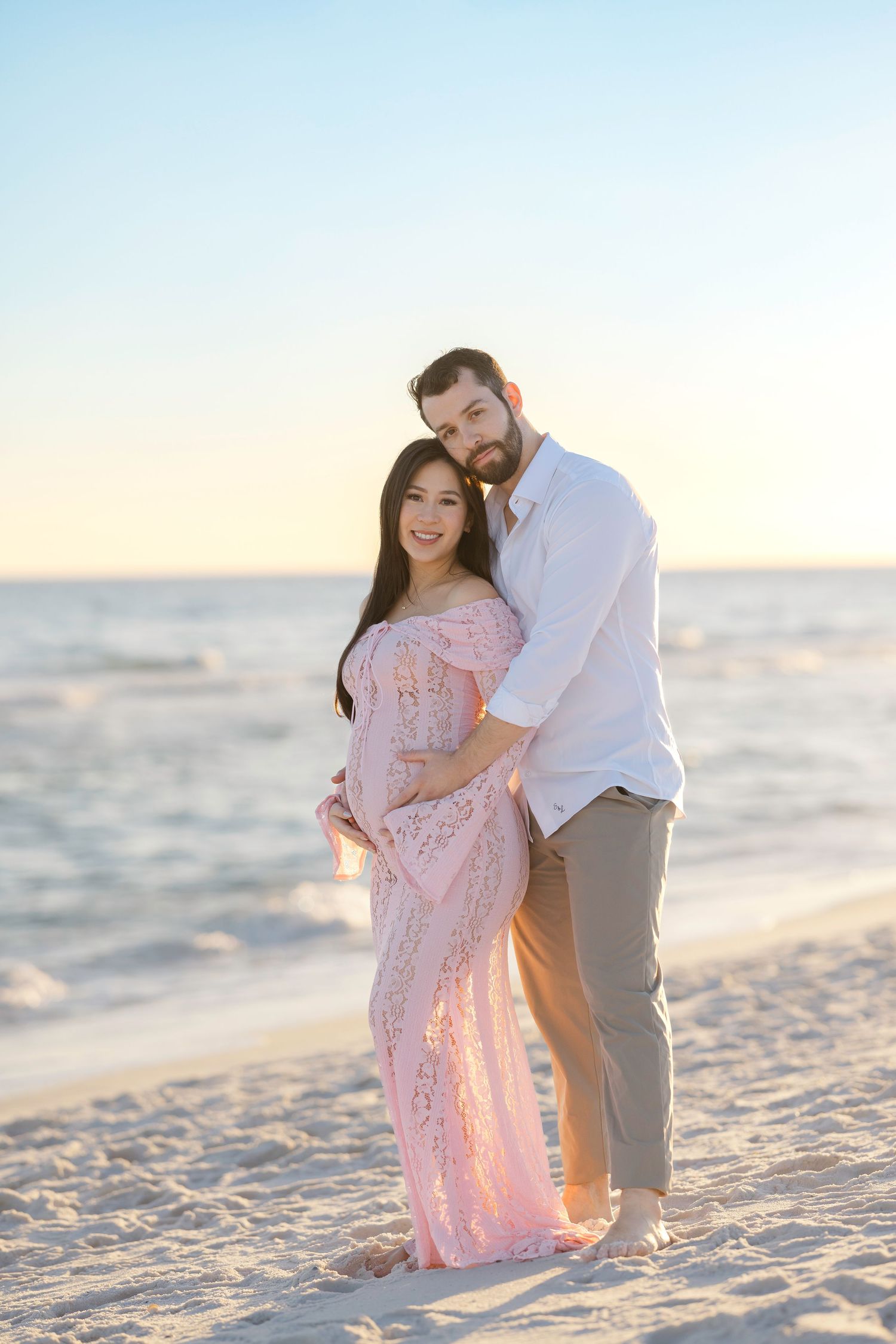 Couple poses on sandy beach at sunset, pregnant woman in pink lace gown embraced by man in white shirt.