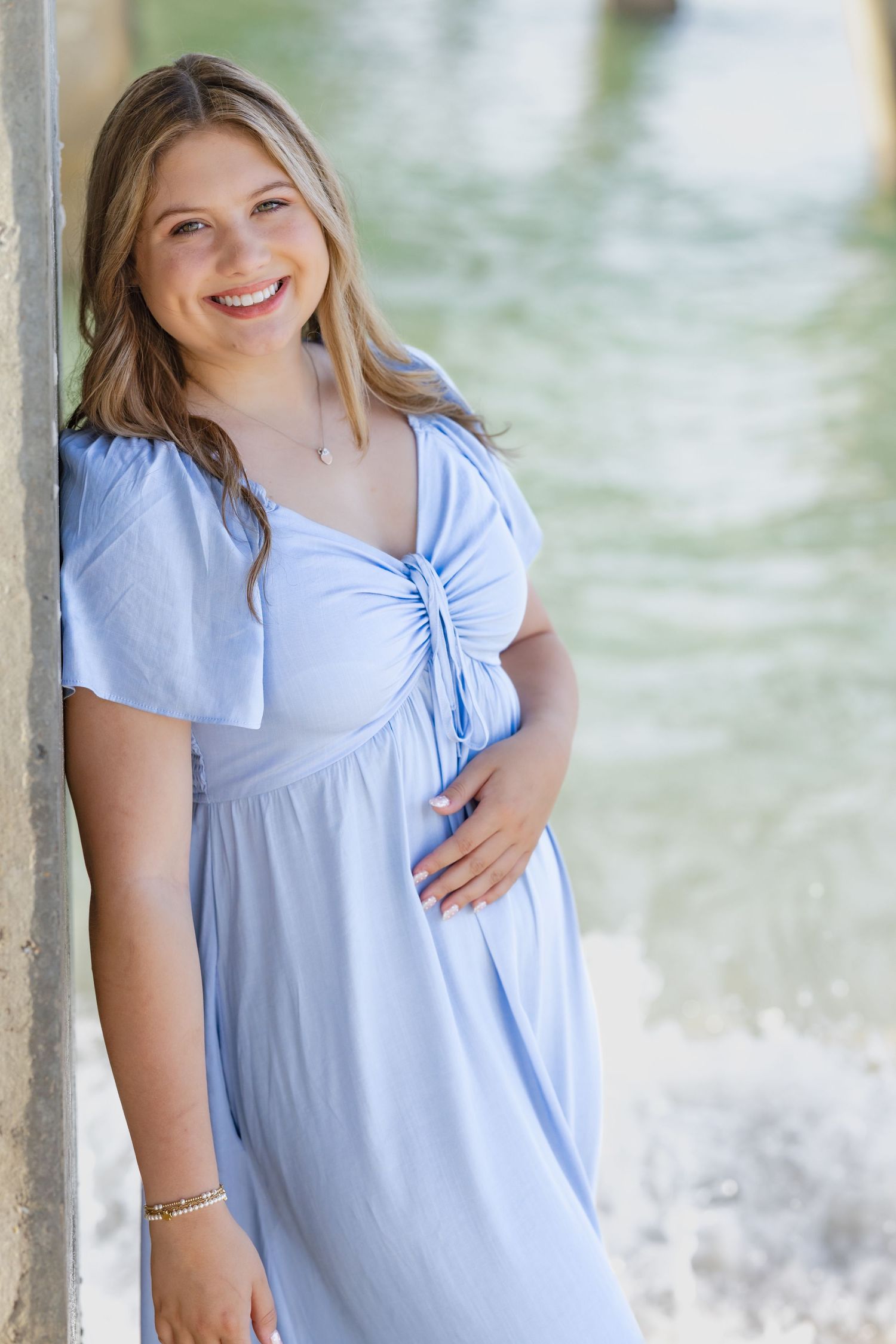 Smiling pregnant woman in light blue dress leaning against pillar near water.
