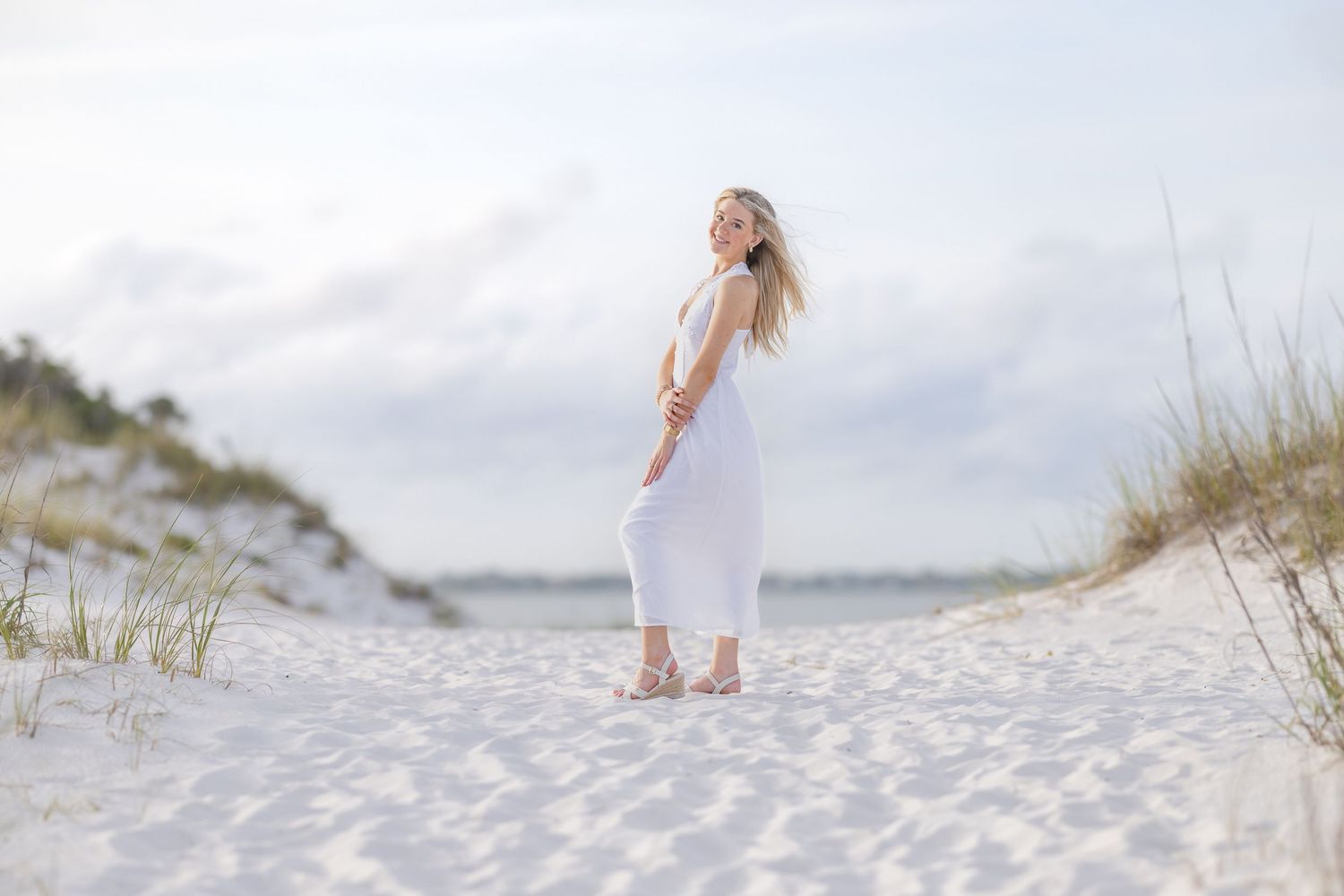 A woman in a white dress stands on a sandy beach path surrounded by dunes and sea grass.