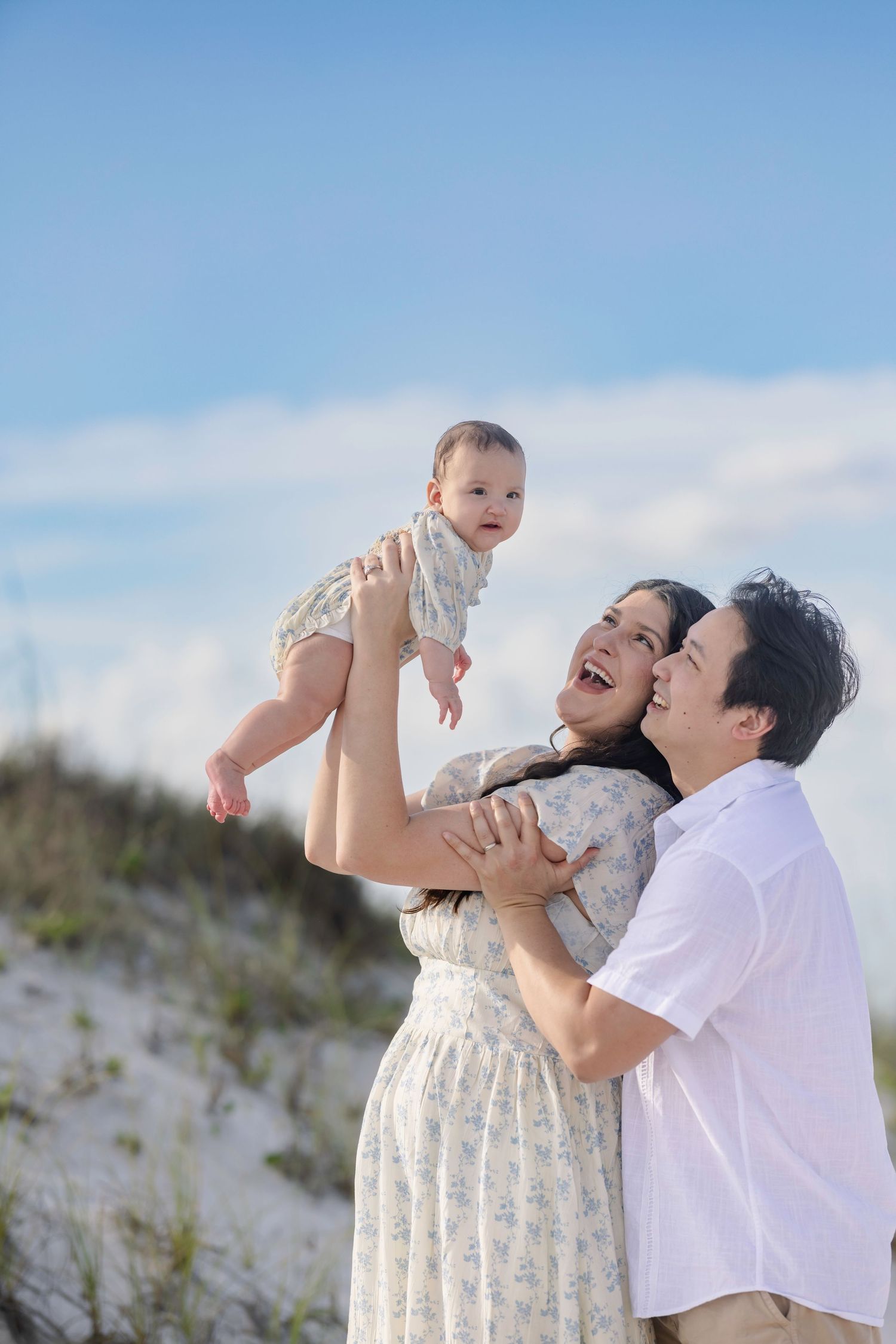 Happy family at the beach, mother lifting baby in the air while father embraces her on a sunny day.