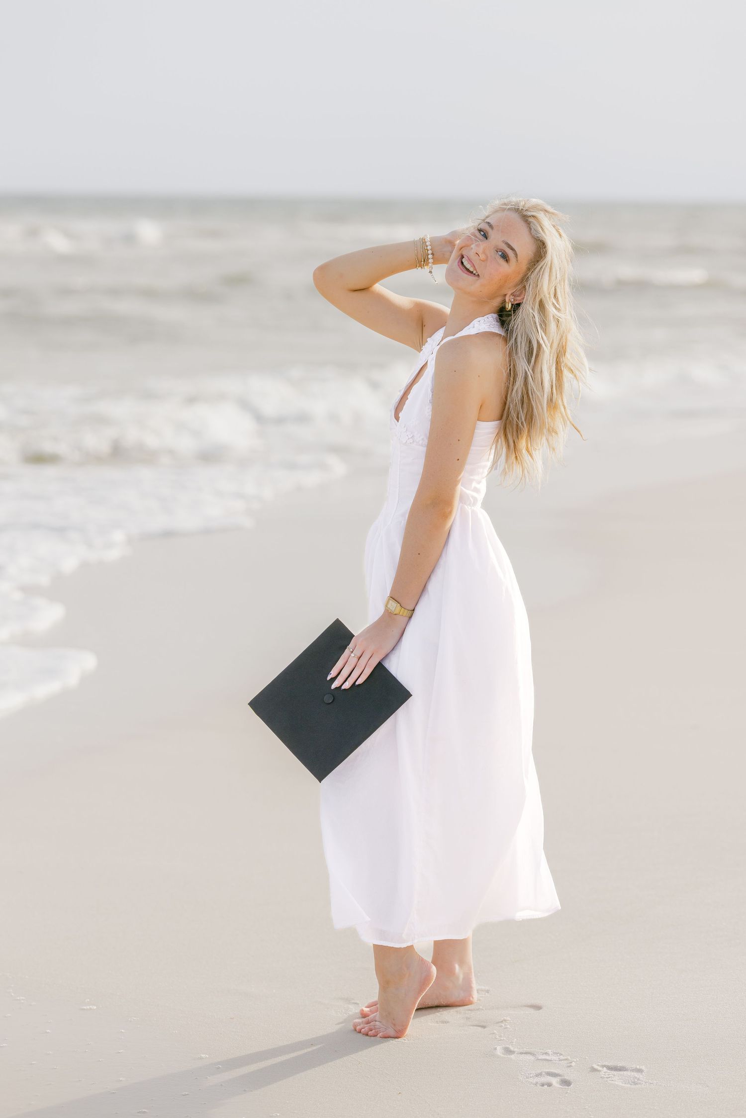 Blonde woman in white maxi dress holding graduation cap while walking on sunny beach shoreline.