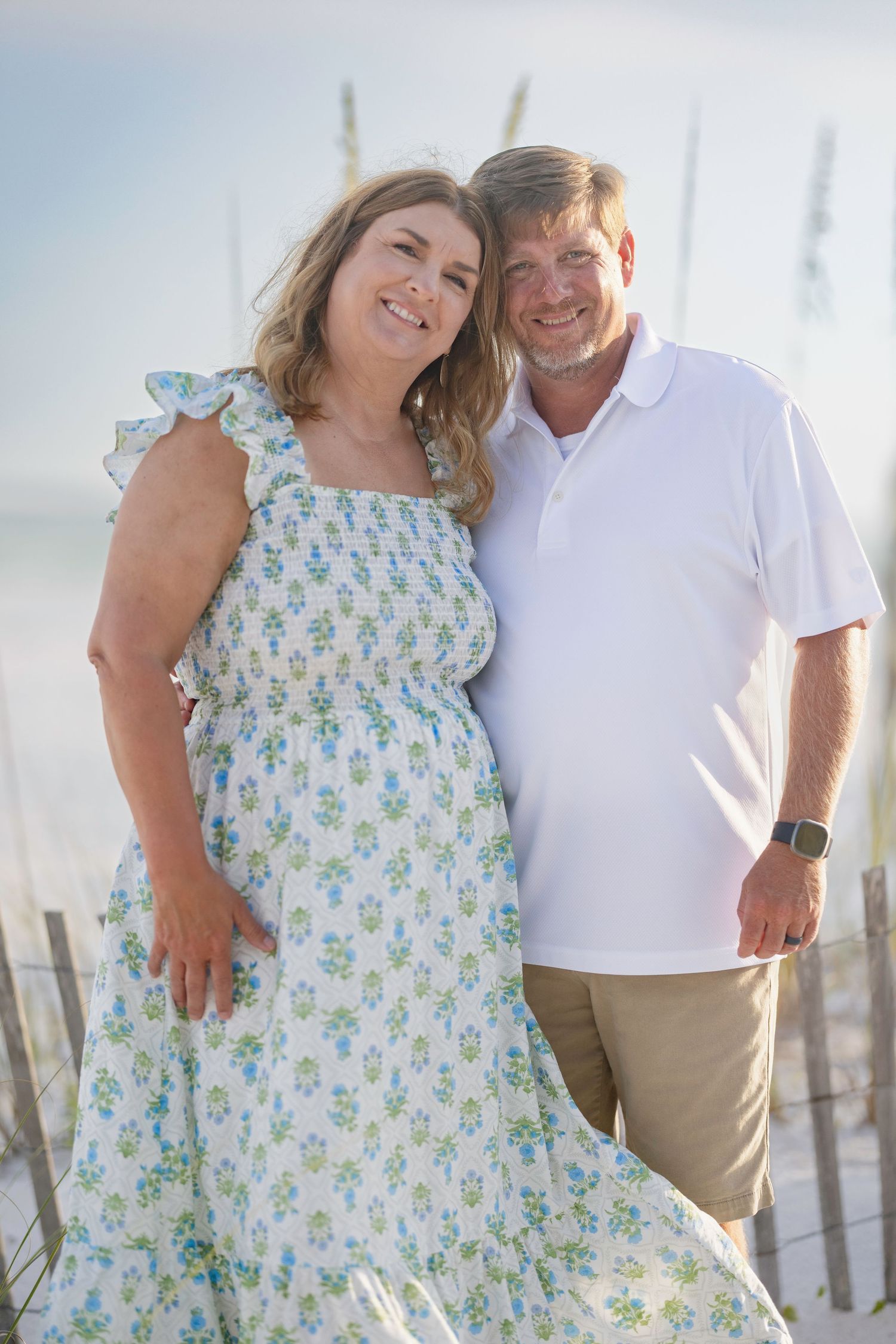 A smiling middle-aged couple poses together on a sandy beach, woman in floral dress and man in white polo shirt.
