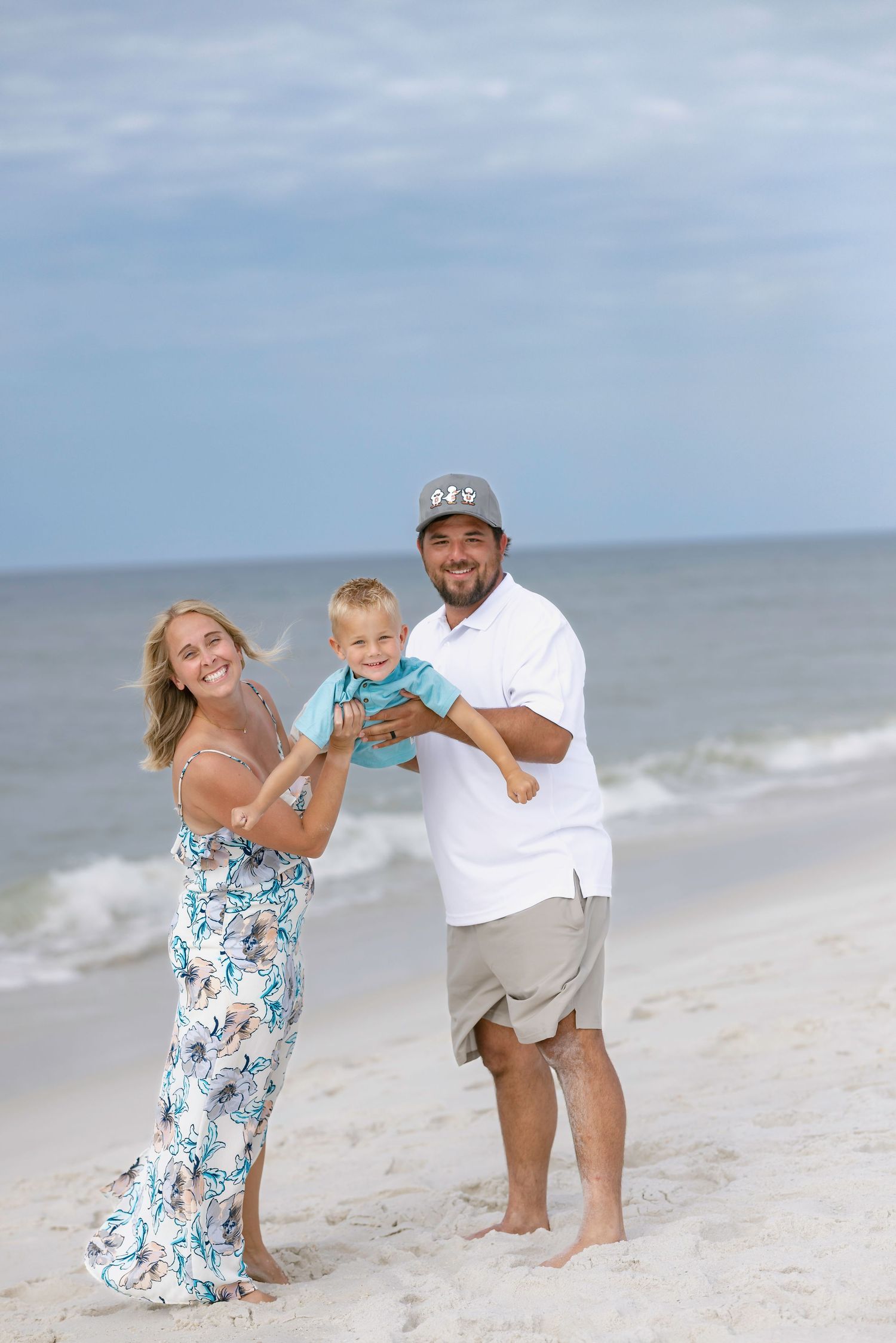Happy family of three posing on a sandy beach, parents holding toddler in turquoise outfit with ocean waves behind.