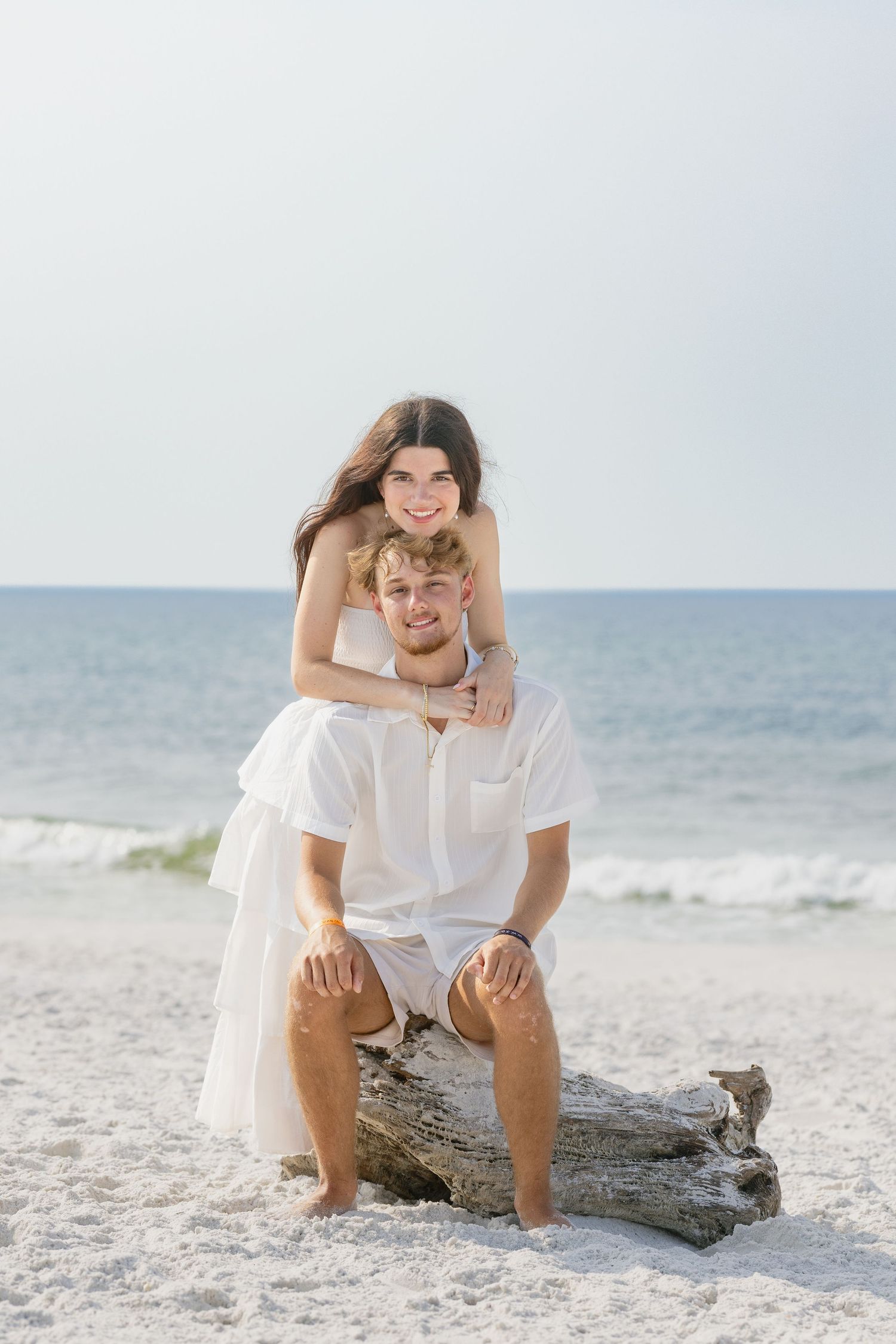 Smiling couple in white outfits posing on driftwood log on sunny beach with ocean behind.
