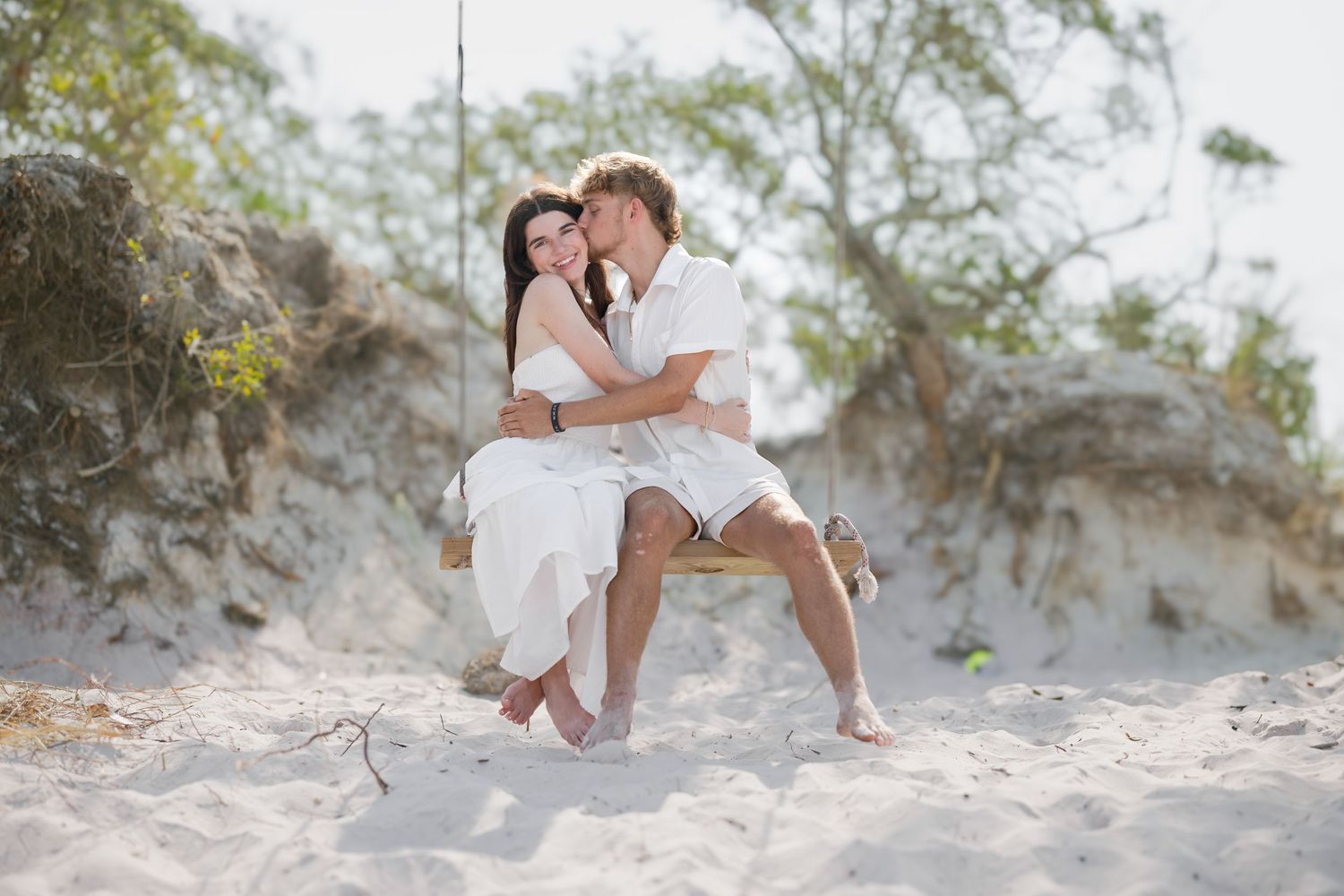 Romantic couple sitting together on a rope swing in sandy beach dunes surrounded by trees.