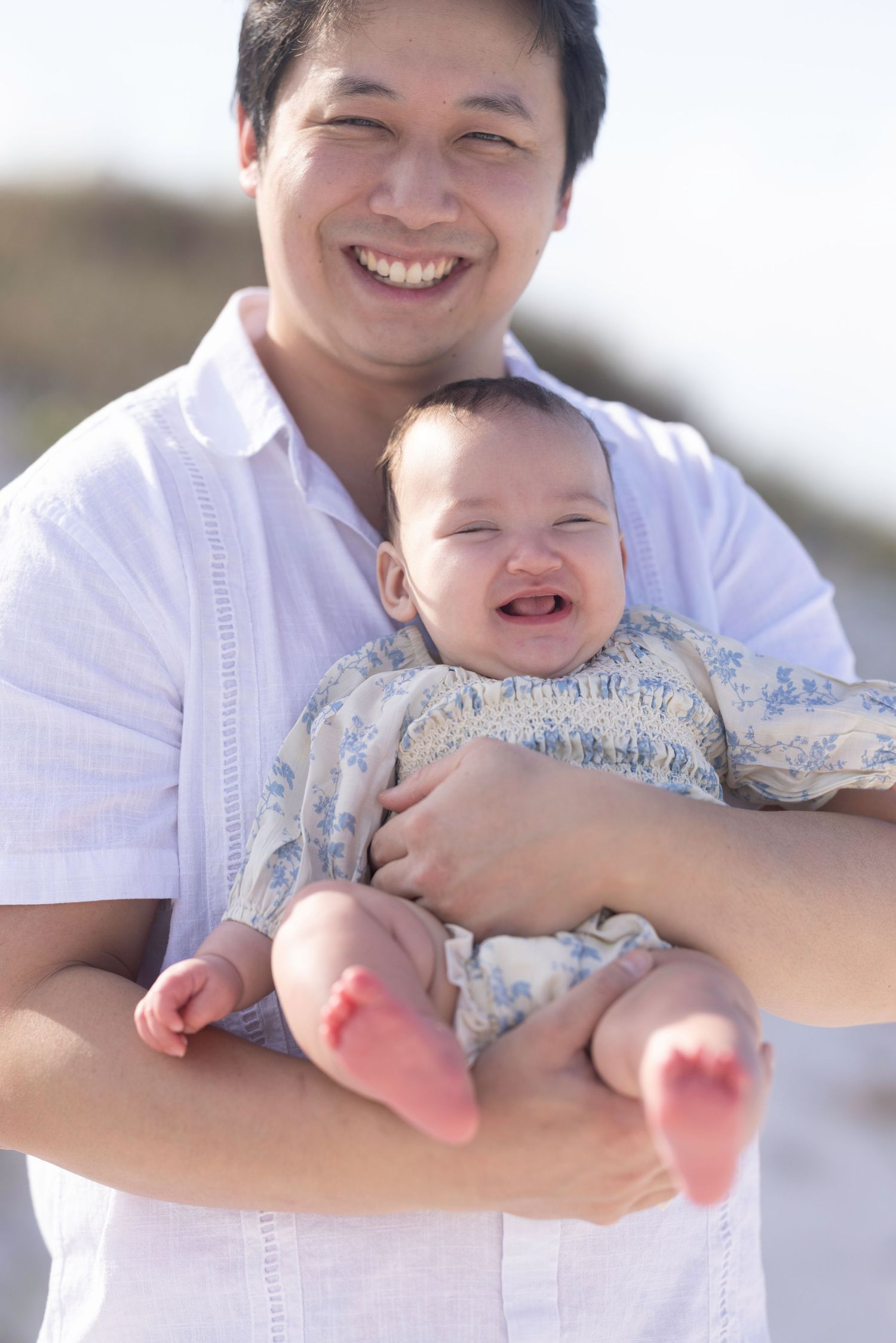 Smiling Asian father holding happy laughing baby outdoors at the beach on a sunny day.