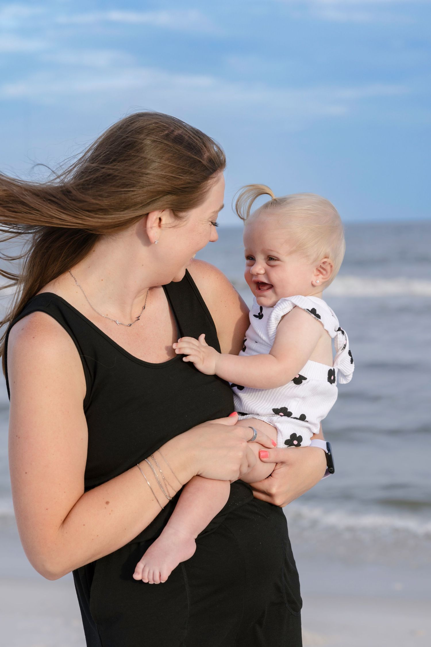 Mother holding laughing baby on a breezy beach, both wearing black and white outfits.