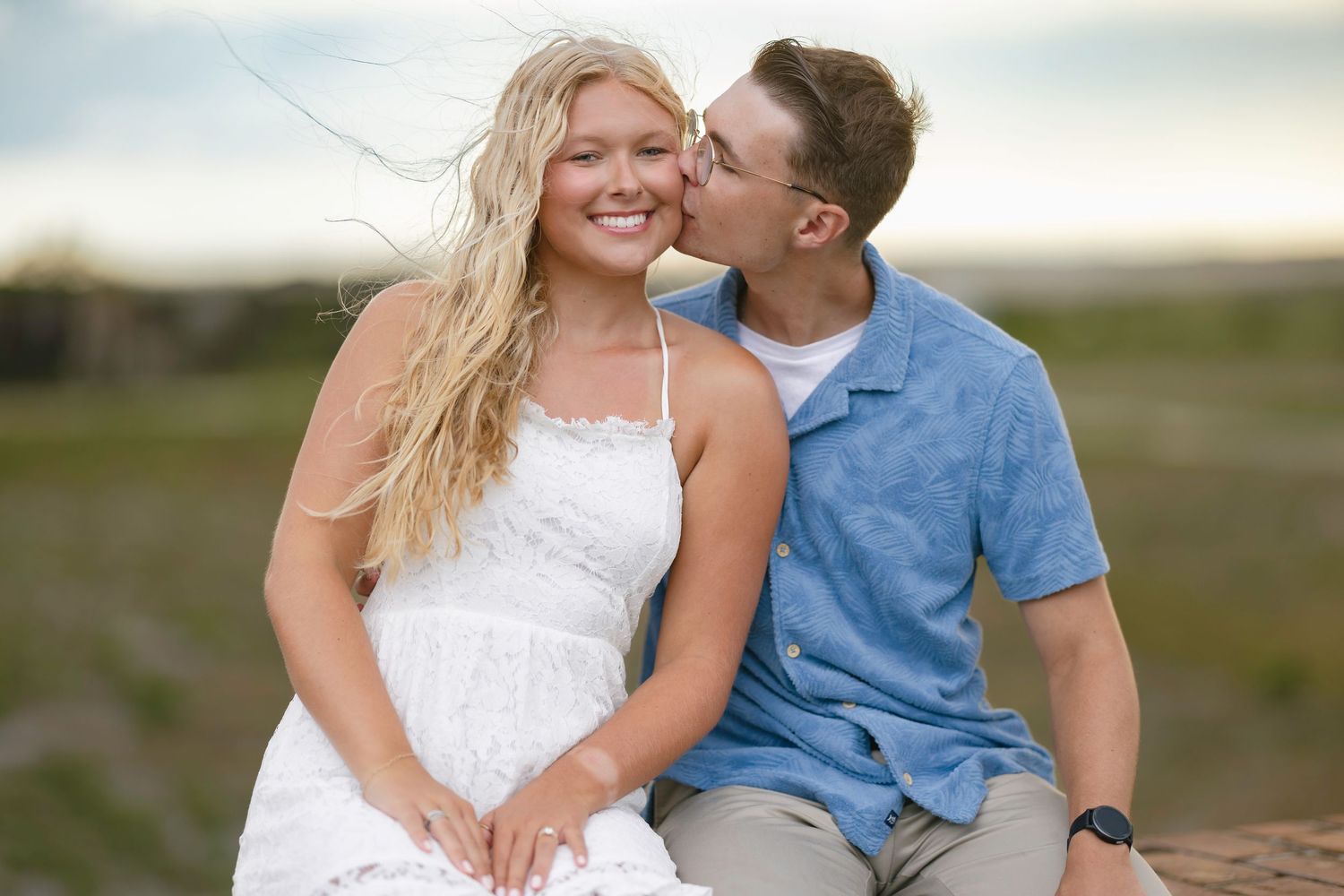 A young couple sharing a romantic kiss outdoors, woman in white dress with flowing blonde hair, man in blue shirt.