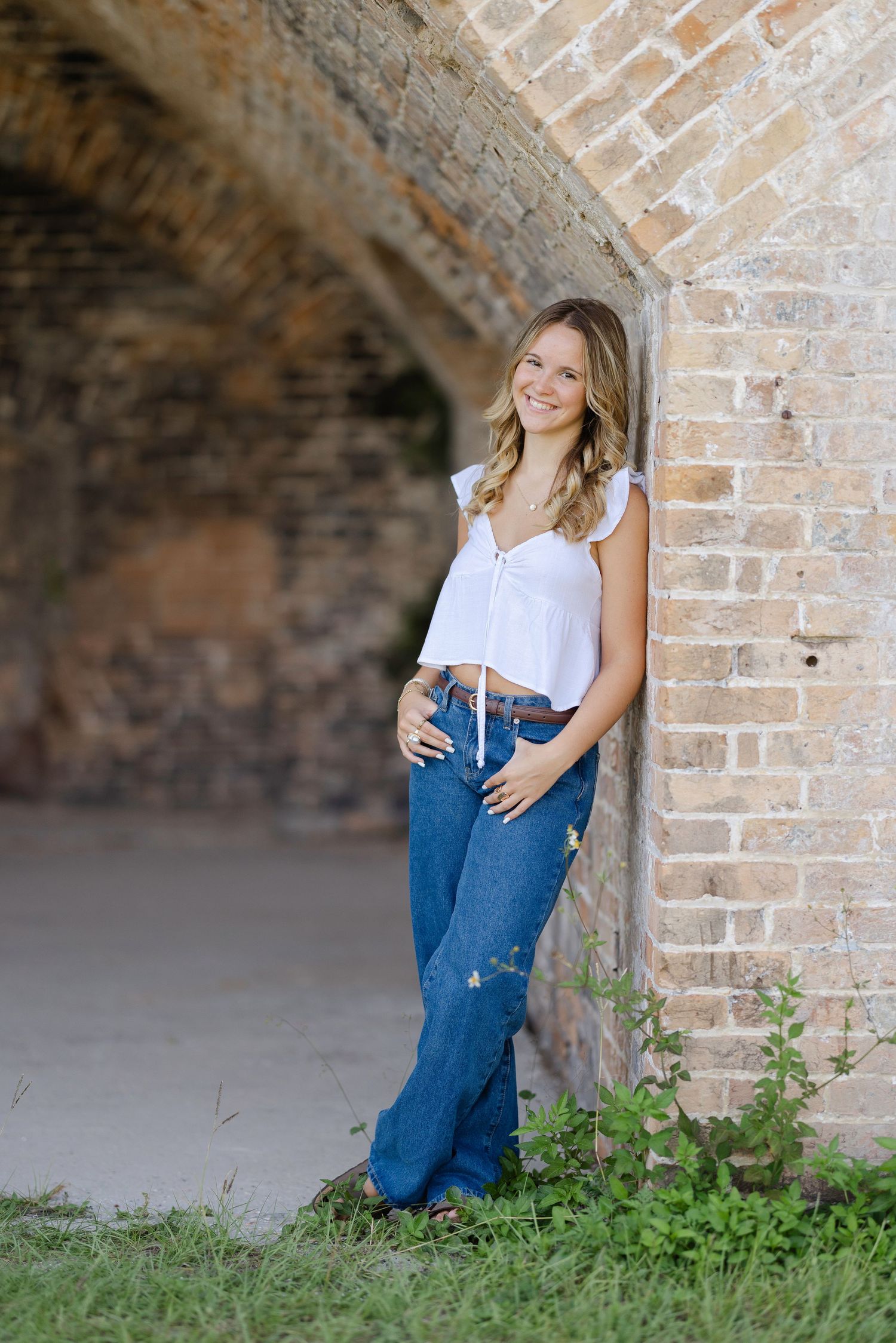 A person in a white sleeveless top and blue jeans poses against a rustic brick wall with greenery at the base.
