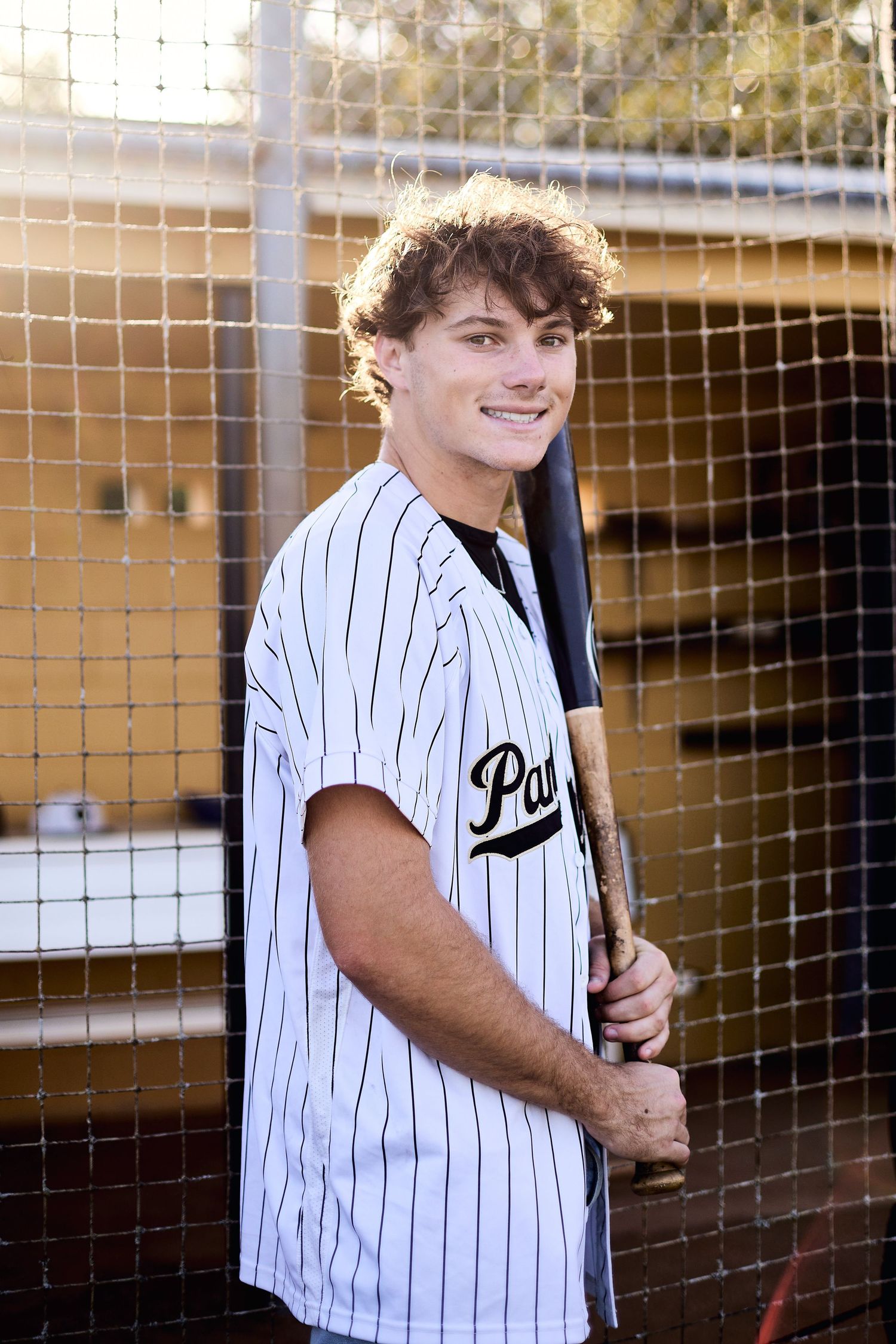 Baseball player in pinstriped uniform smiles while holding bat near dugout netting during golden hour lighting.
