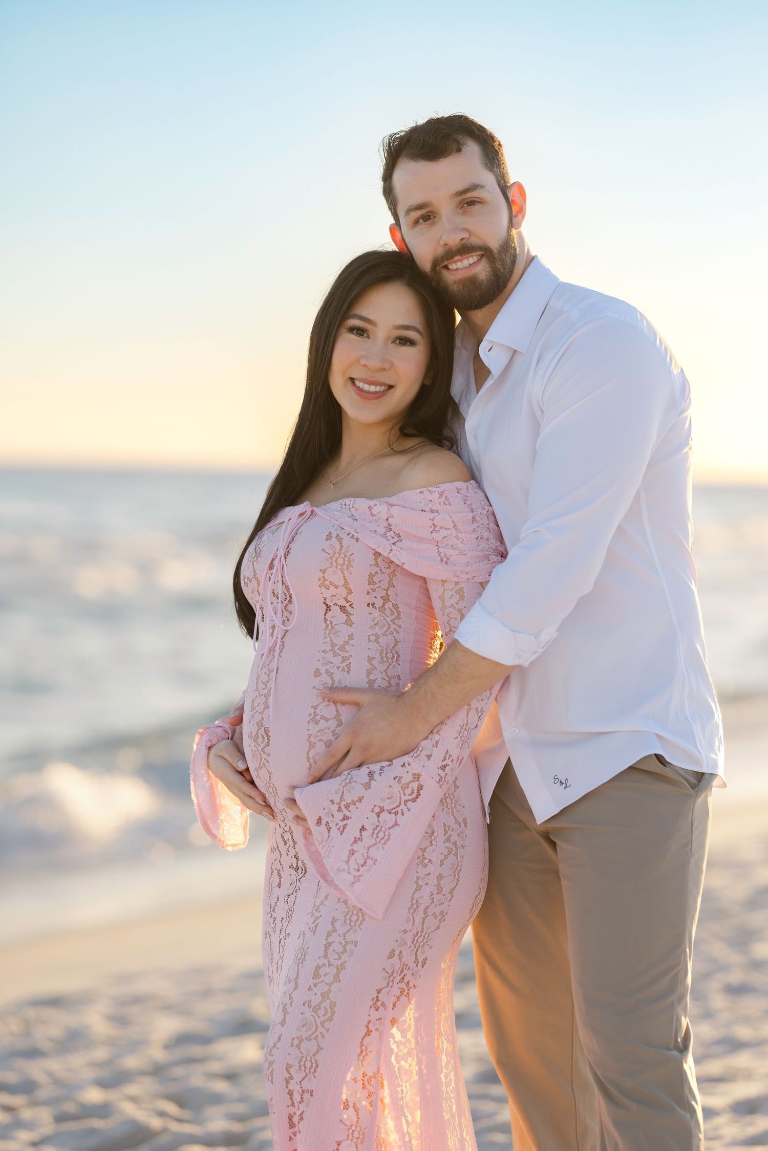 A couple embraces on a white sandy beach during sunset wearing pink and white outfits.
