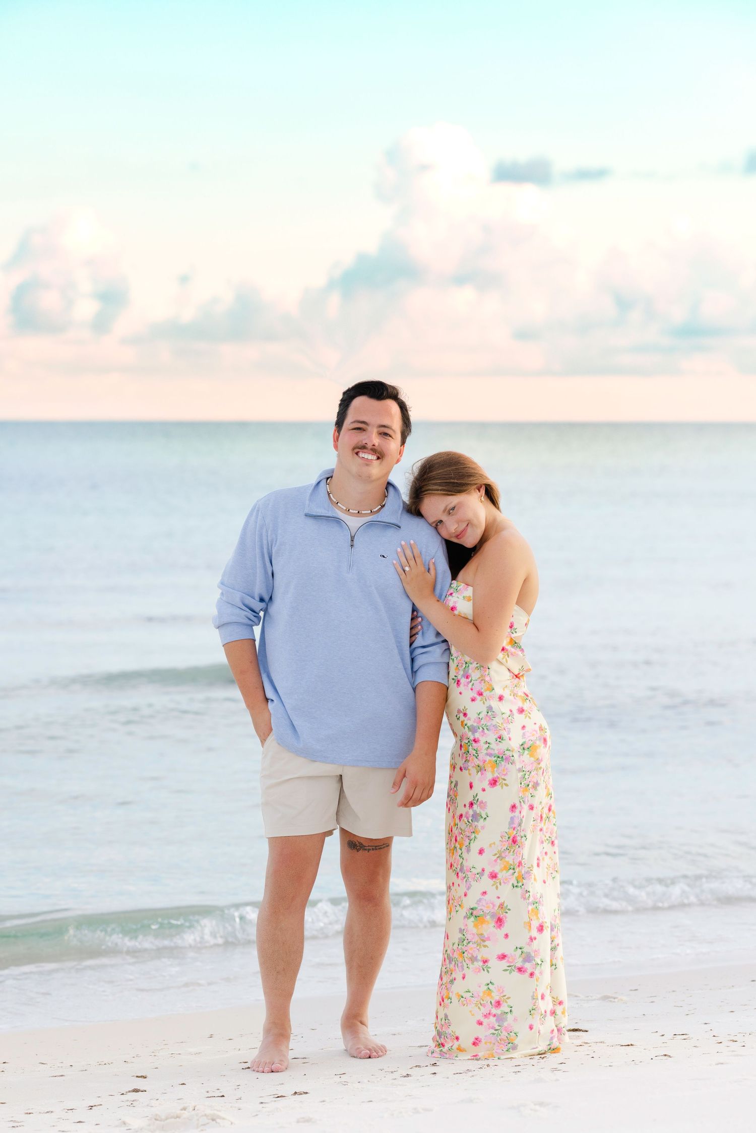 A couple shares a tender moment while standing barefoot on a peaceful beach at sunset with soft pastel clouds in the sky.