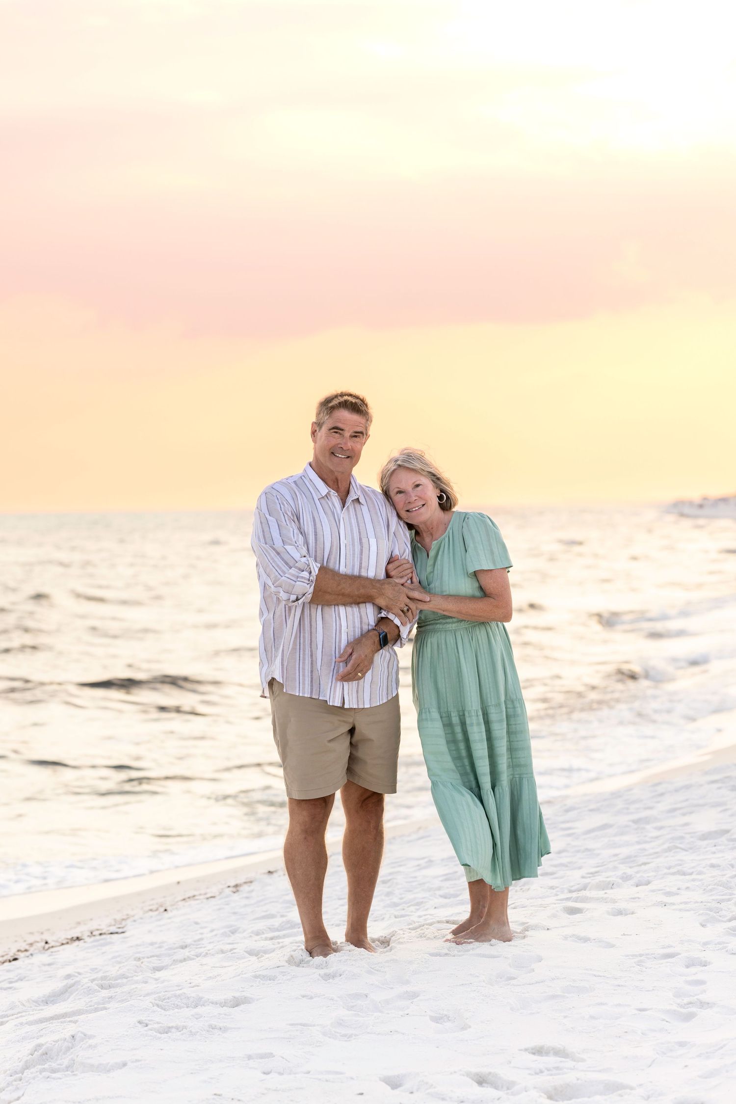 A couple shares a romantic moment while walking along a beach at sunset wearing pastel colored clothing.