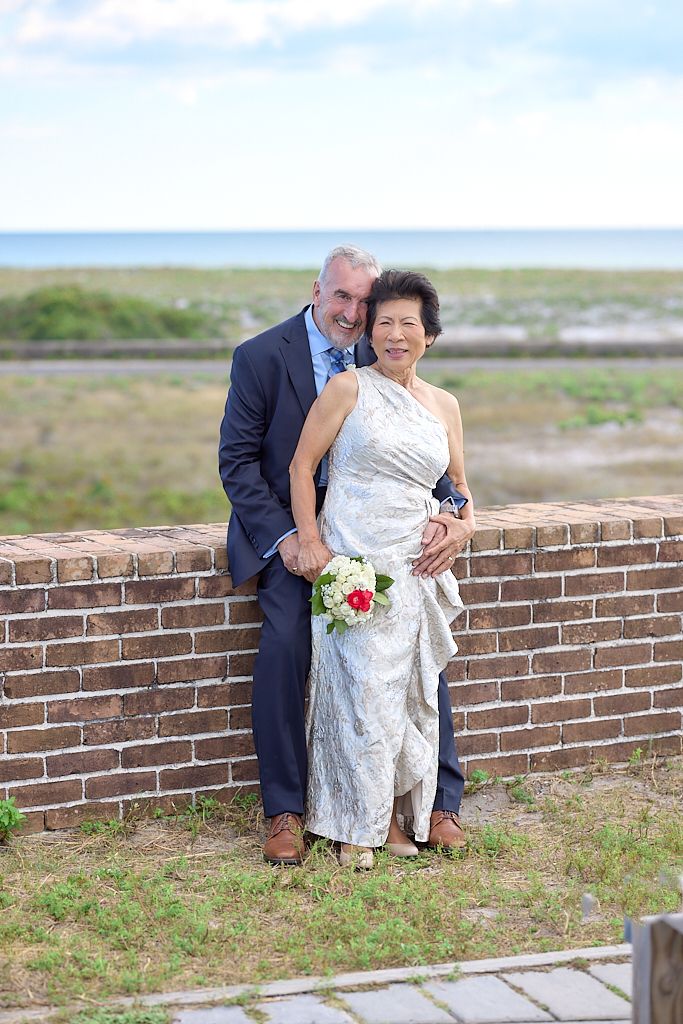 Couple in wedding attire sits together on brick wall overlooking scenic coastal landscape.