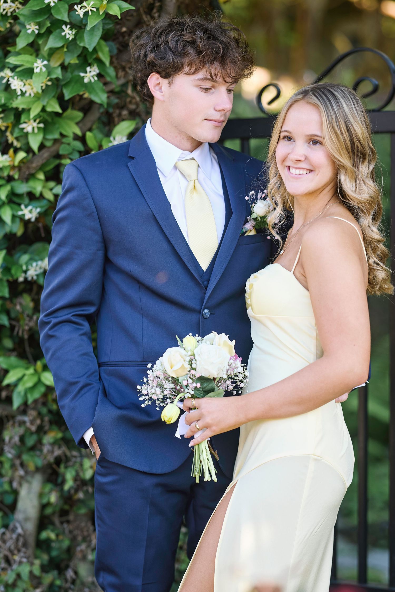 A couple in formal wedding attire poses together outdoors with greenery and white flowers in the background.