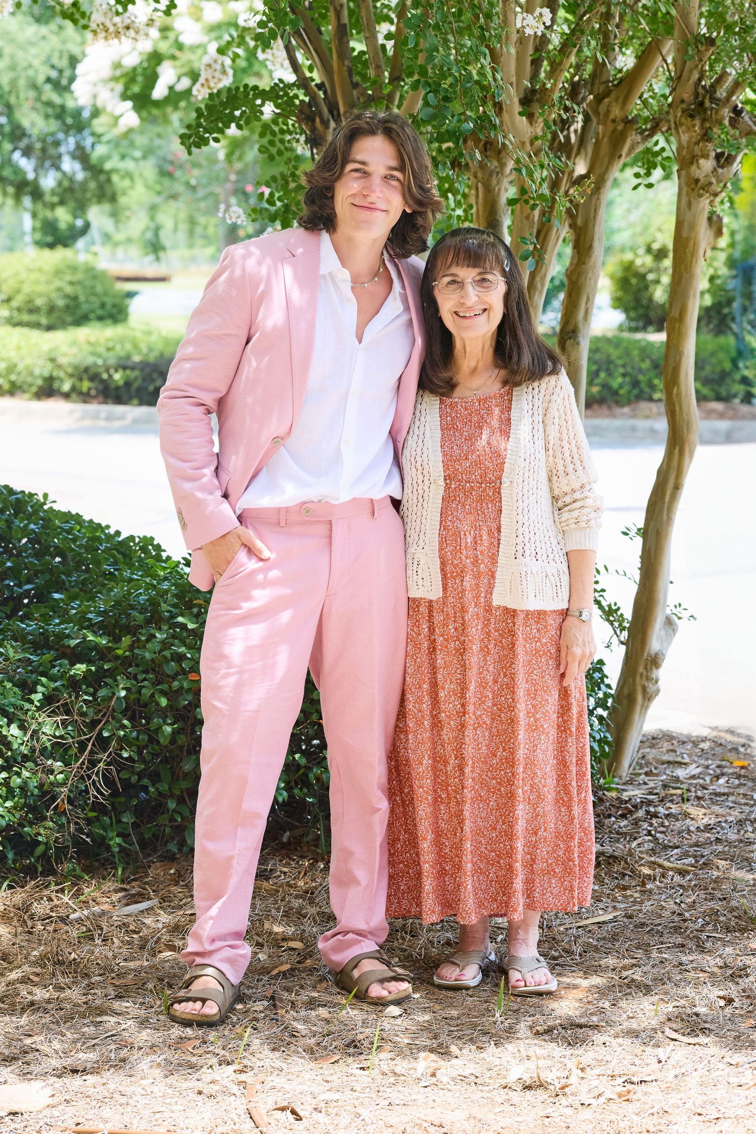 A couple poses outdoors, with one wearing a pink suit and the other in a flowy peach dress.
